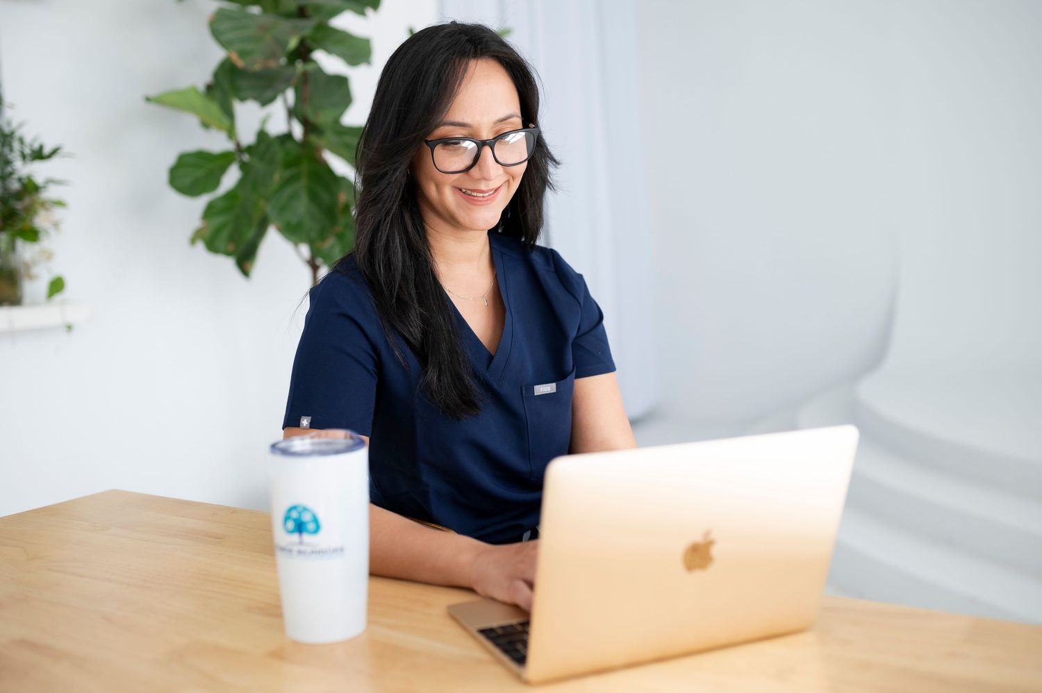 Woman in navy shirt working on MacBook laptop in bright modern office setting.