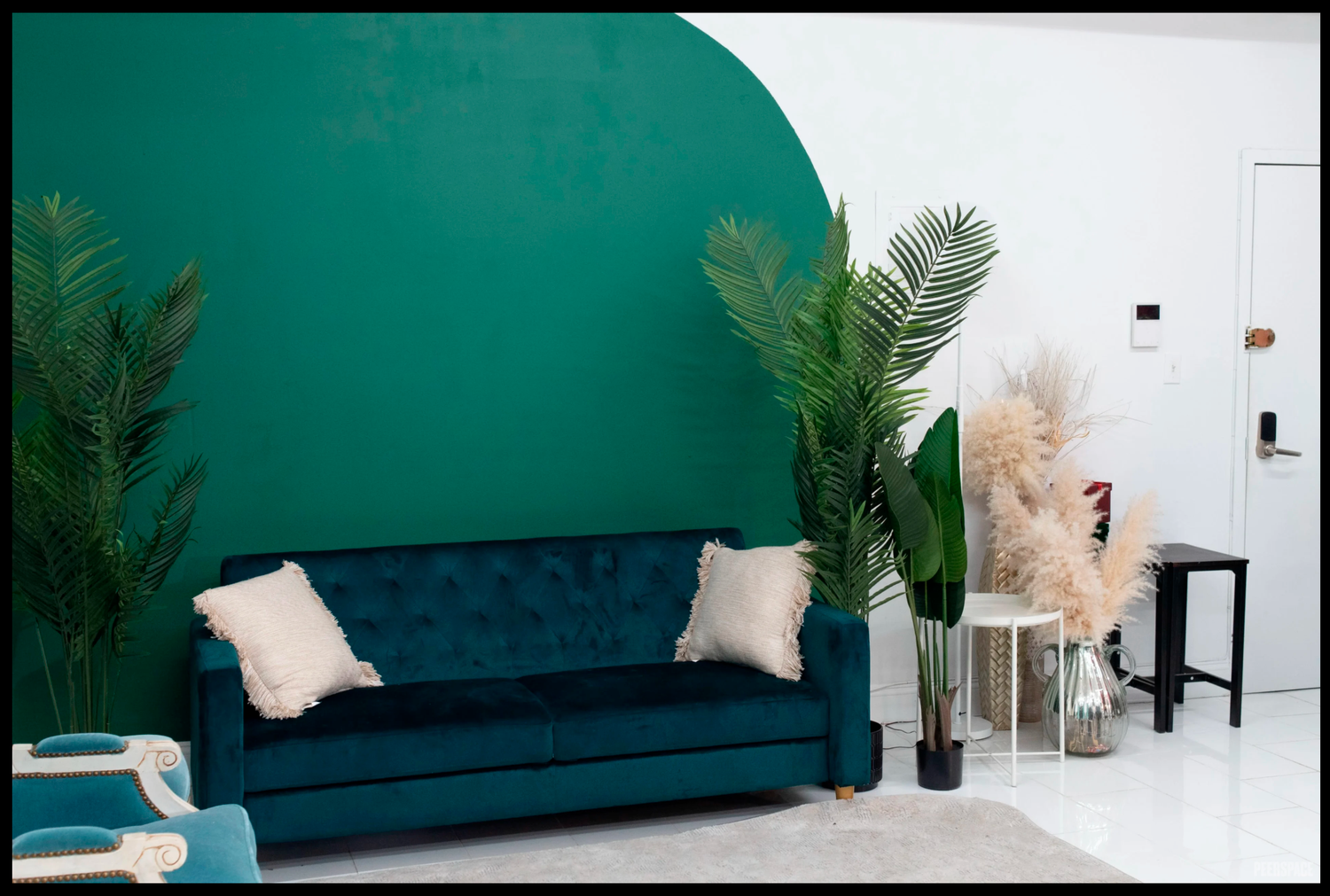 Modern living room with teal velvet sofa, pink cushions, green arch wall, and tropical plants on white floor.