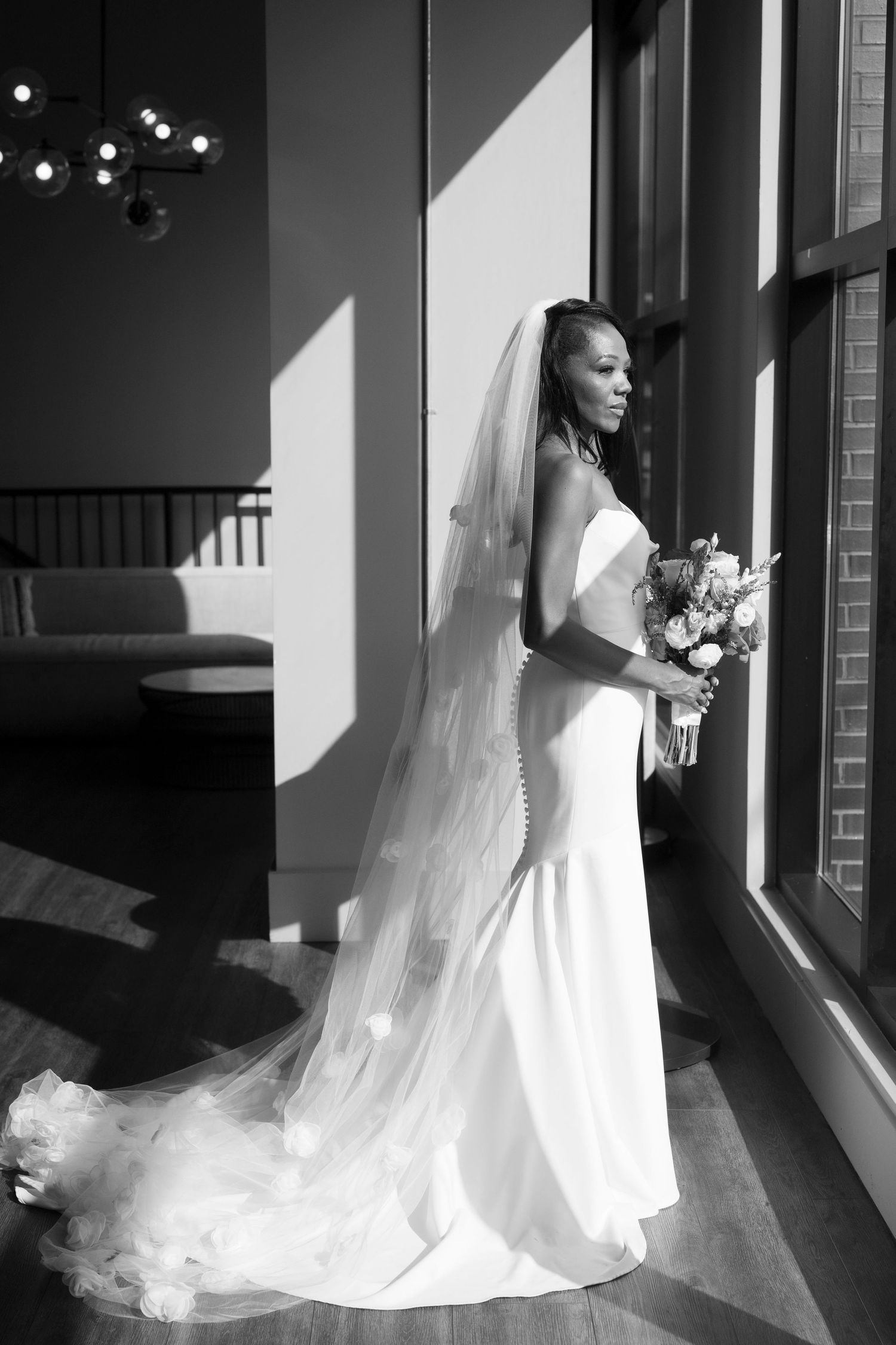 Black and white photo of a bride in a white gown and veil holding a bouquet while gazing out a sunlit window.