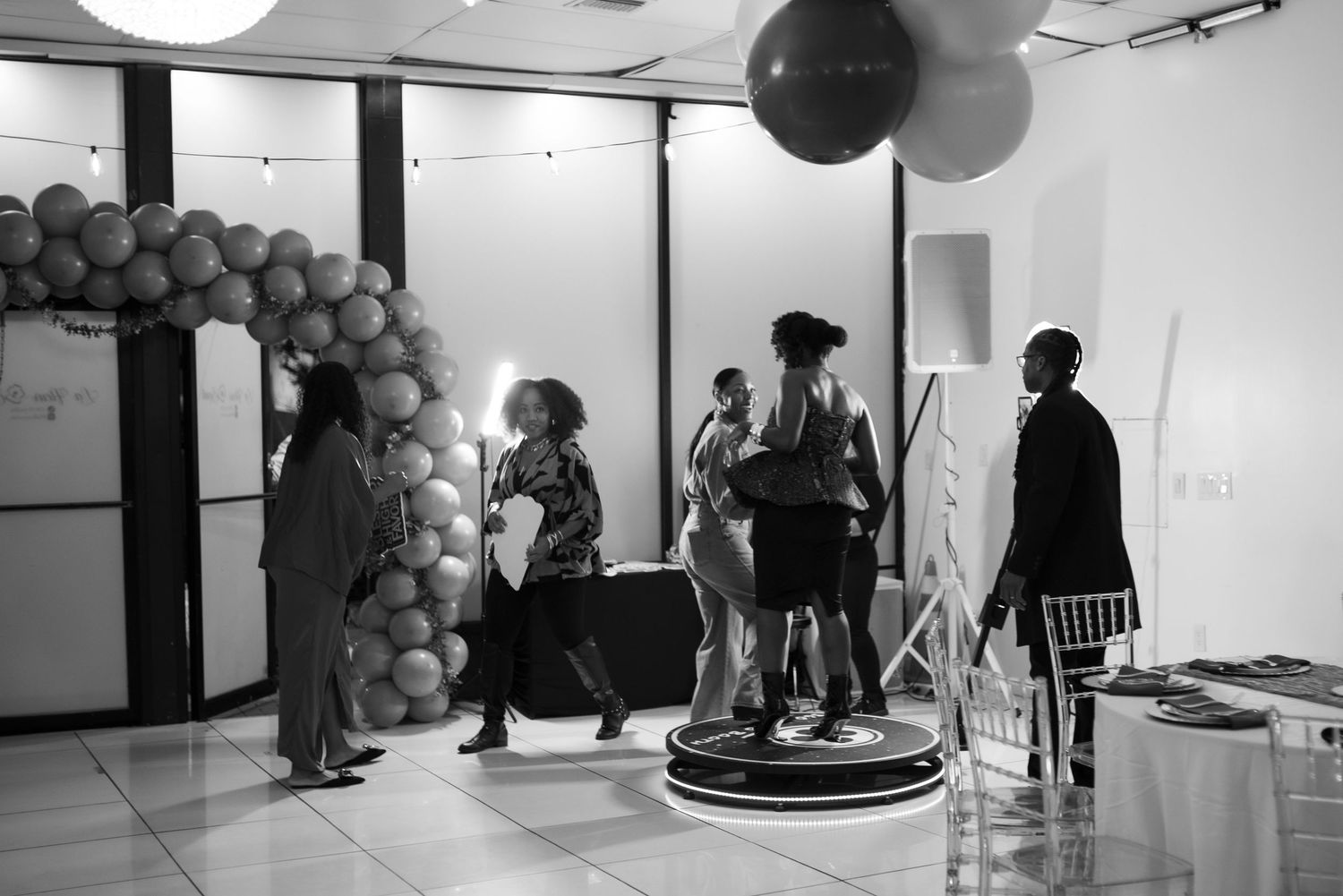 Black and white photo of guests mingling at an indoor event with balloon arch decor and a photo booth.