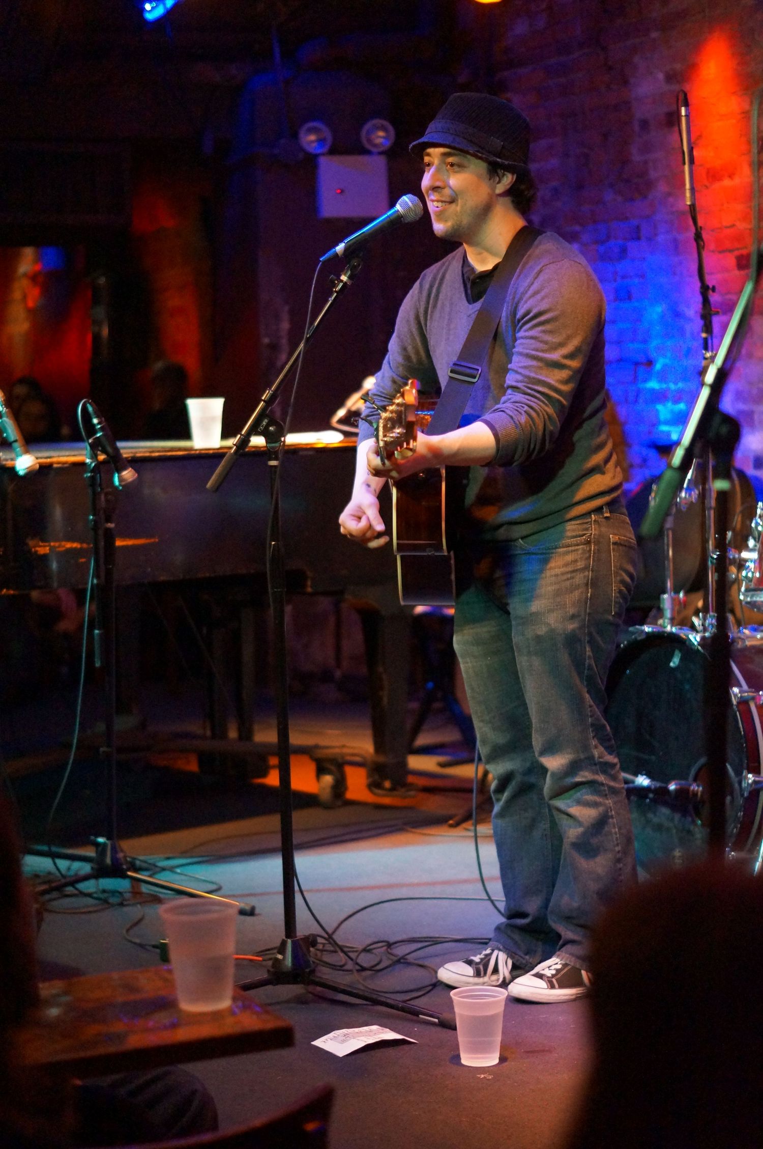 Male guitarist performing on stage with microphone, wearing beanie hat in dimly lit music venue.