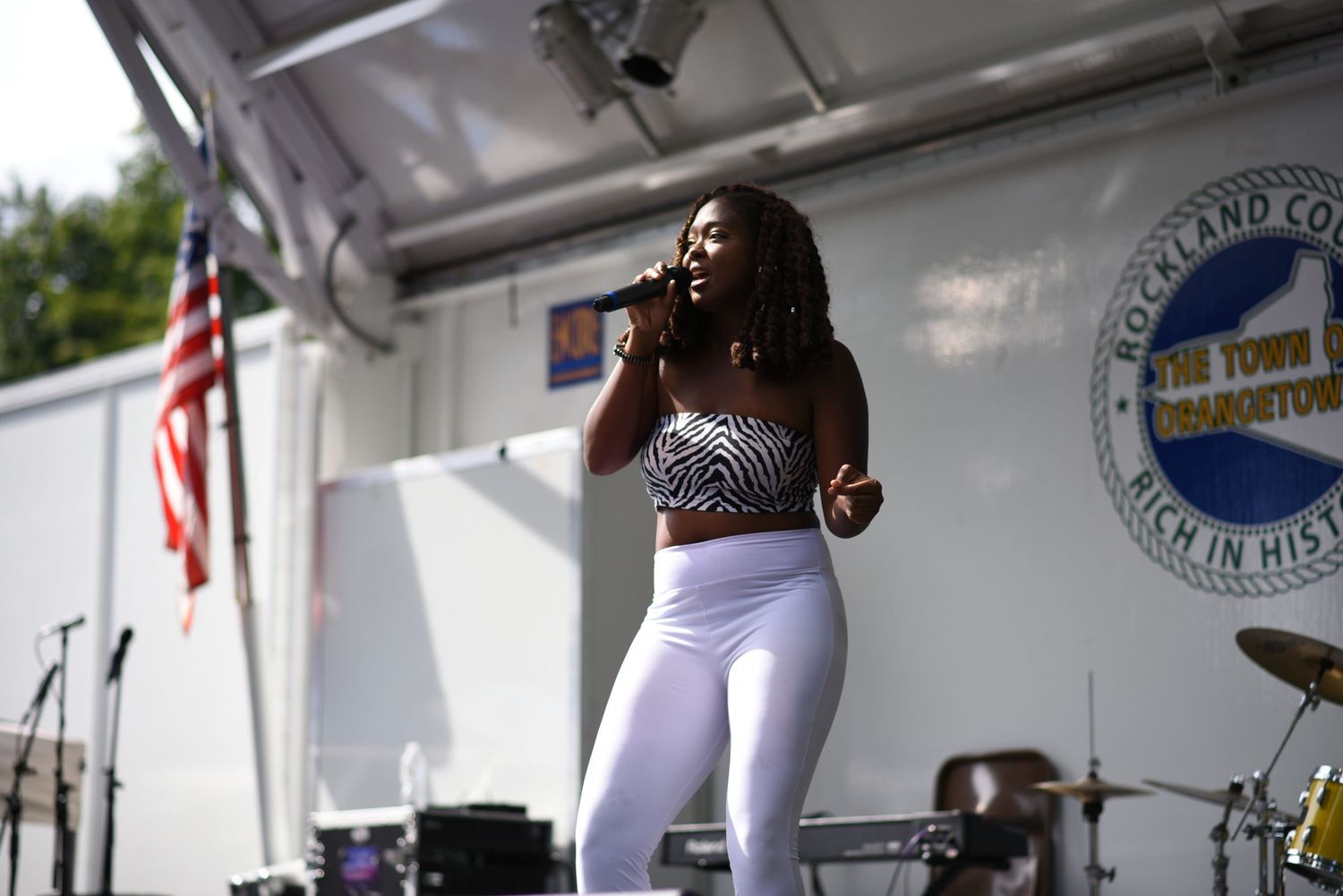 Young woman singing on outdoor stage wearing white leggings and zebra print top at Rockland County town event.