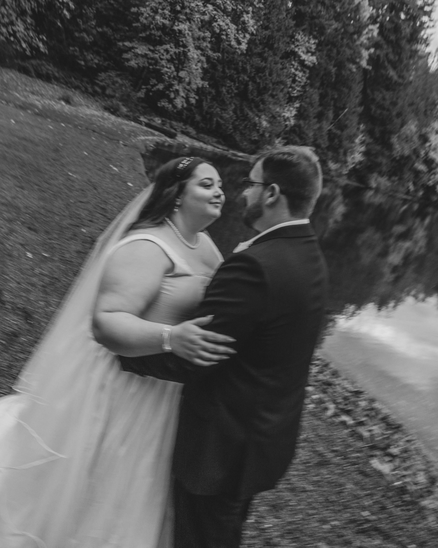 Black and white photo of couple standing together beside a lake wearing their wedding attire. Grainy, blurred, film inspired.