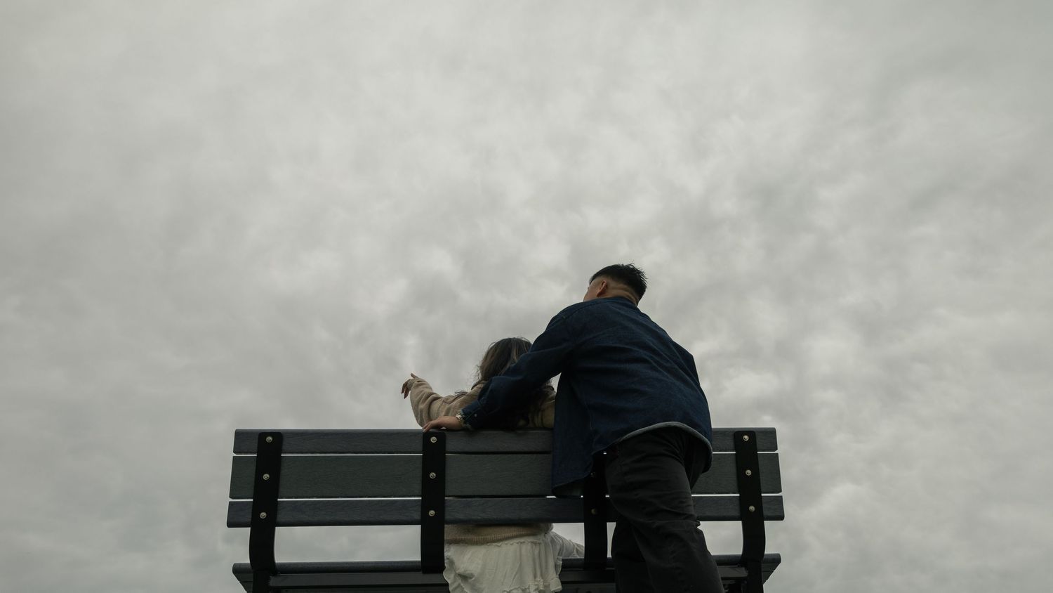 Couple together, woman sitting on a bench, pointing up towards the sky, man standing behind her at the bench. BC engagement photographer