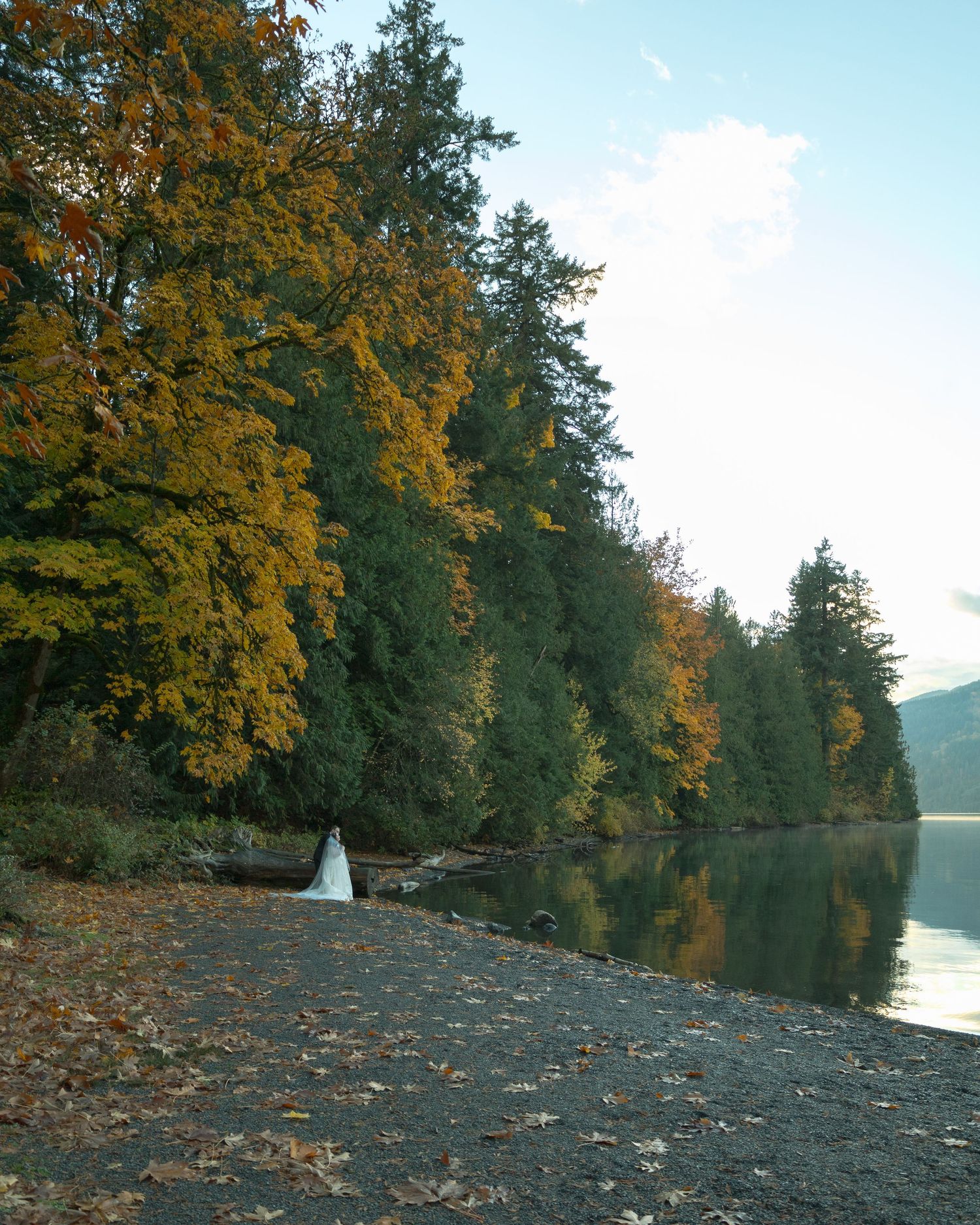 Couple stands together by the edge of a lake, woman wearing a flowing white wedding dress. Photo features fall colours and a nostalgic grain