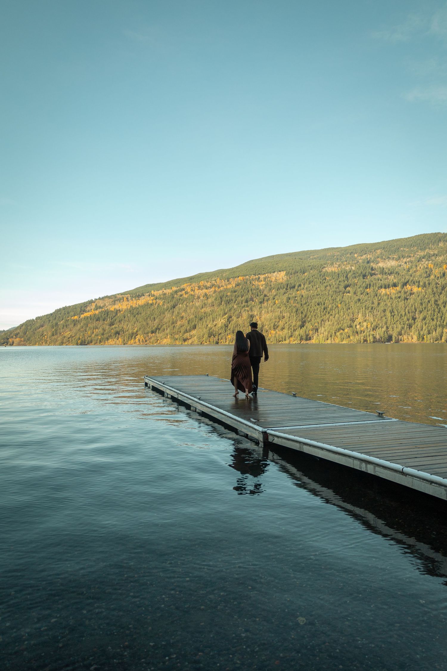 Couple walking together along a dock, lake and mountain in the background. BC engagement photographer