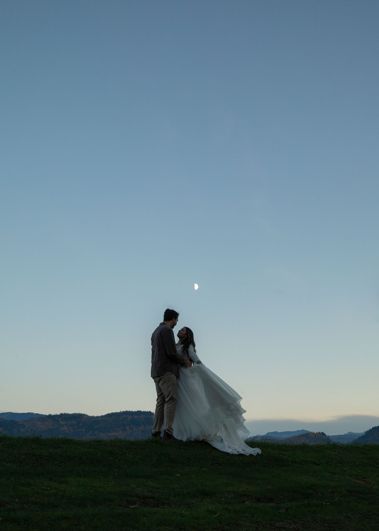 Cinematic style photographer, couple in elopement attire stand against the sky, the moon in the sky above them,