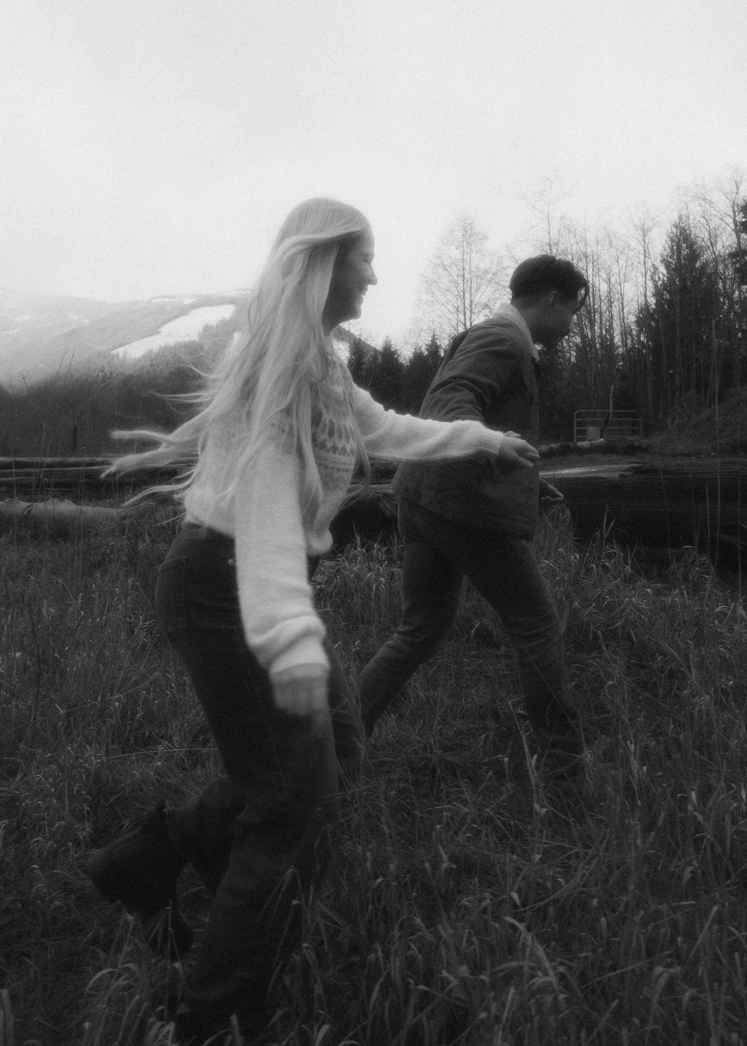 Black and white photo, couple running through grass. mountain backdrop, holding hands. Motion blue, hair blowing in the wind. Grainy, film