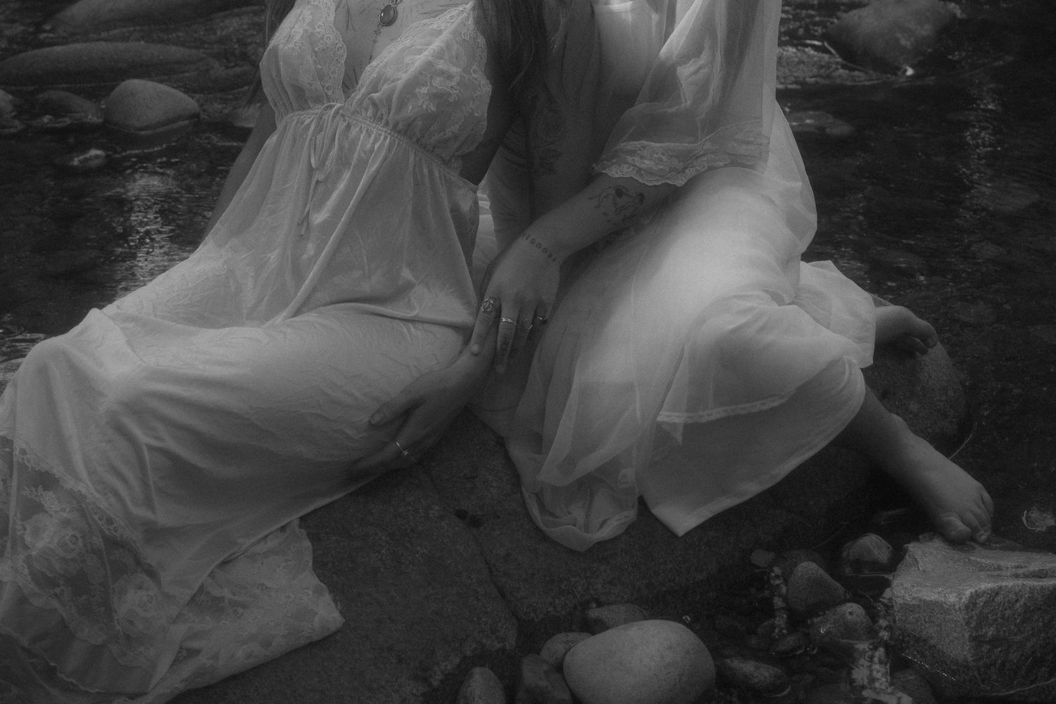 black and white image of women sitting on rocks at a stream, grainy, film inspired, wearing white flowing dresses