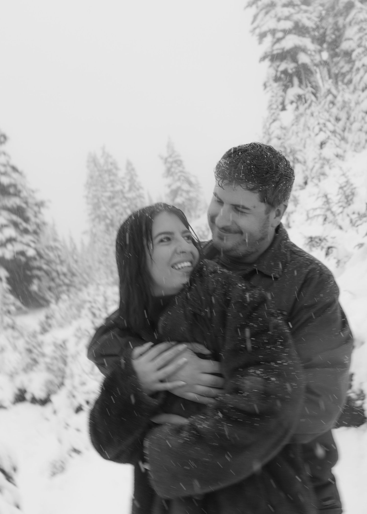 black and white photo of couple, man embracing woman from behind in a hug, woman looking over her shoulder at him smiling. Snowy background