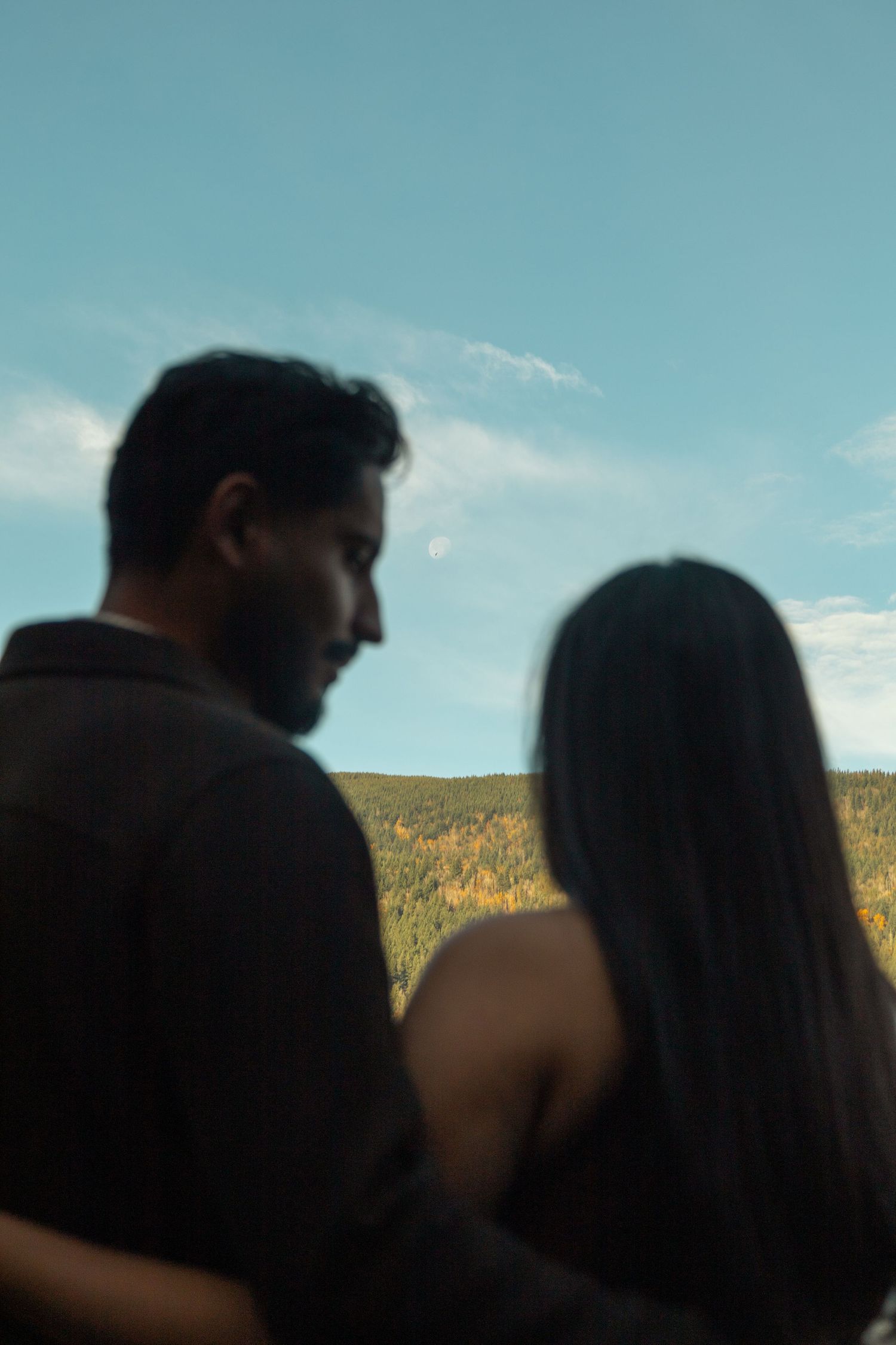 Blurred foreground, couple standing together looking up at the moon that is in focus.