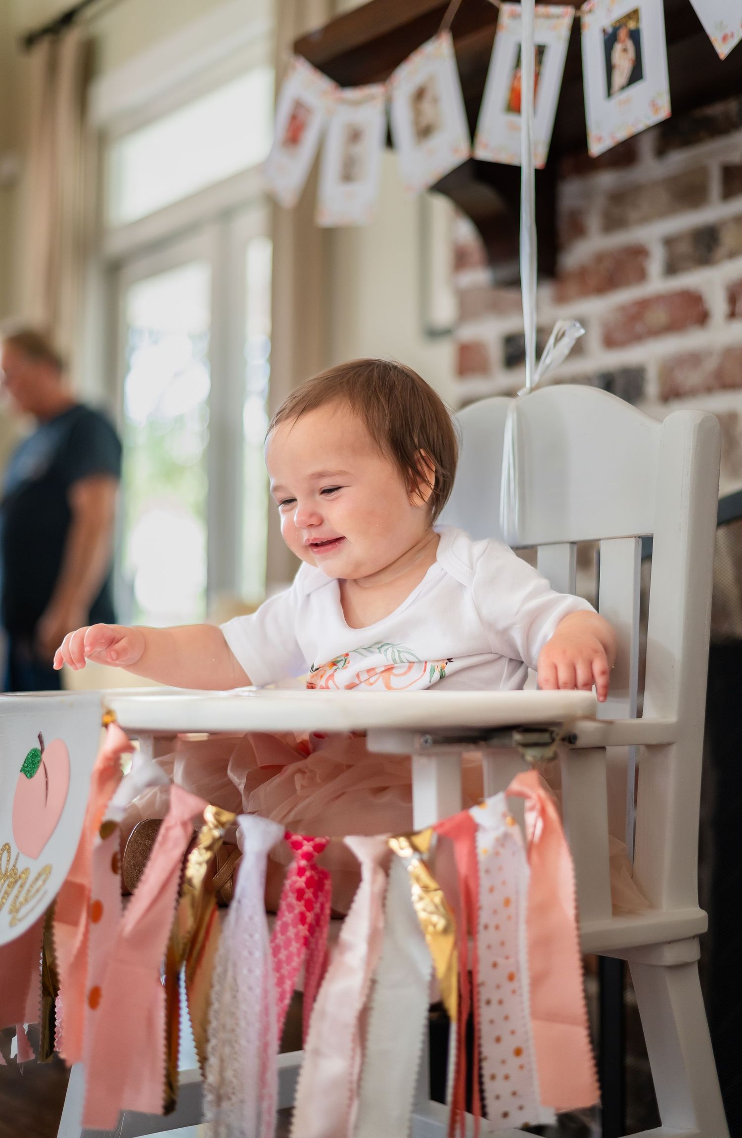 A baby sits at a table decorated with pink and white bunting banners during a celebration.