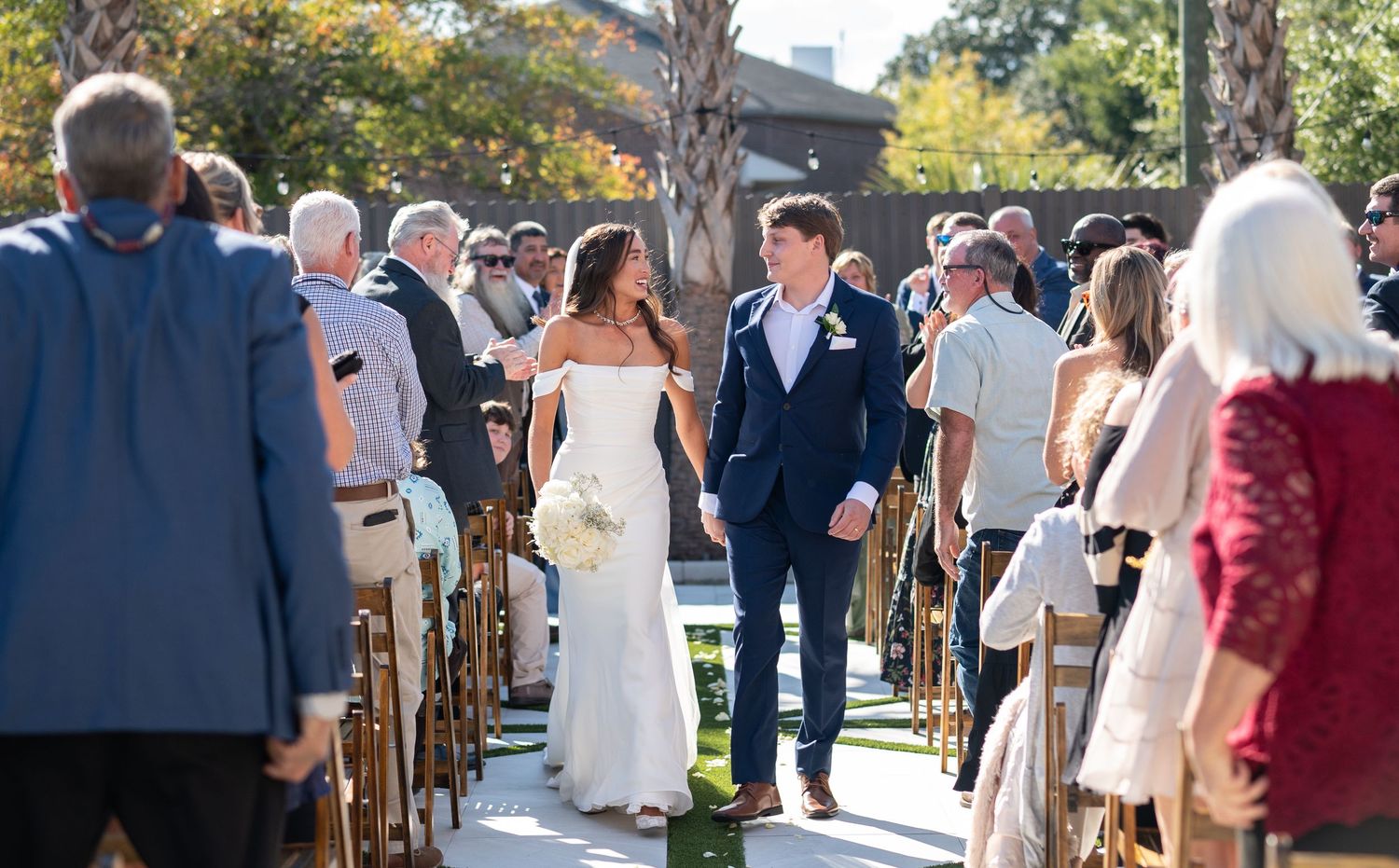 Wedding ceremony guests seated outdoors under trees on a sunny day.