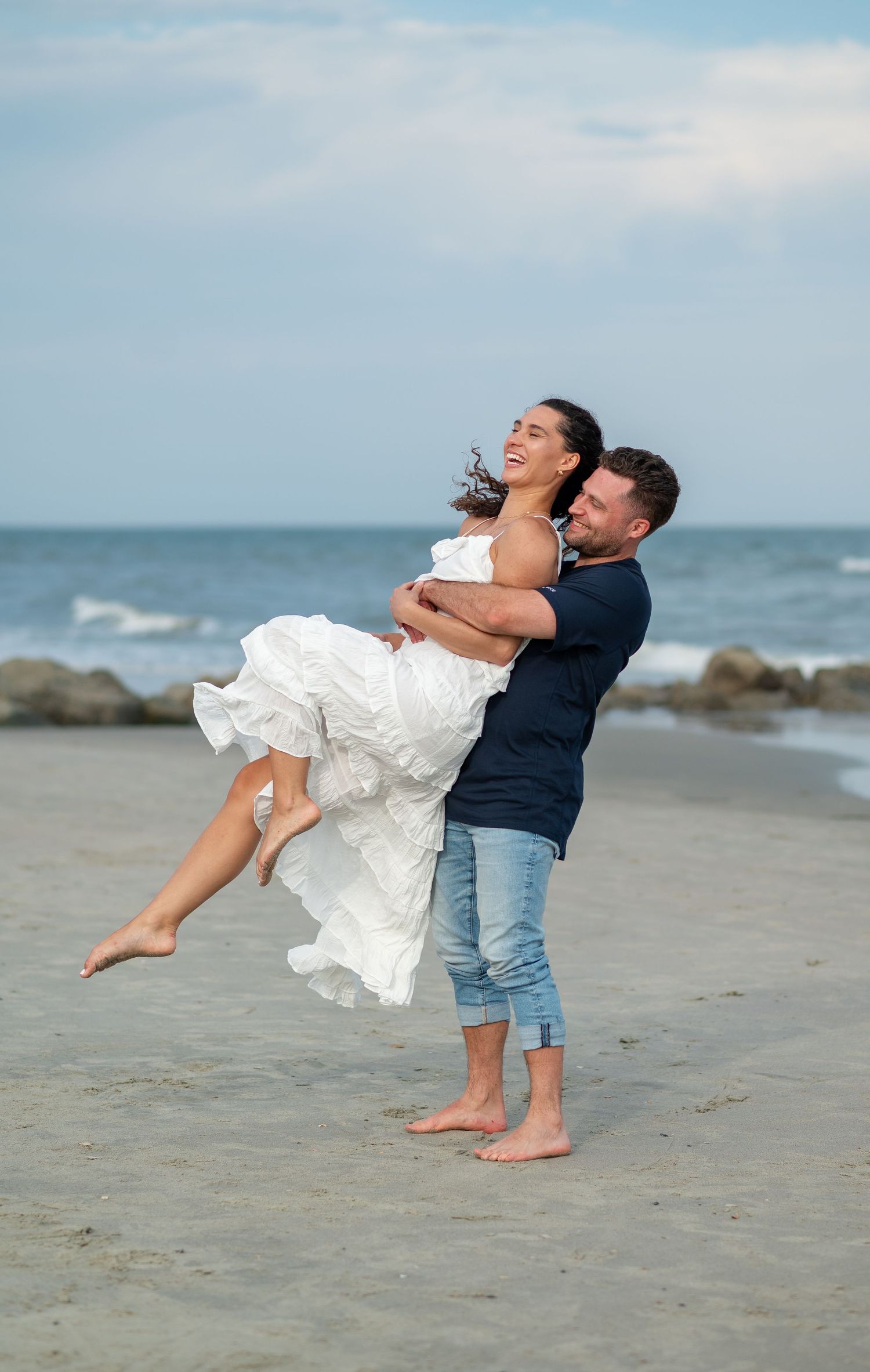 A couple embraces on a beach with waves and rocks in the background as they share a joyful moment together.
