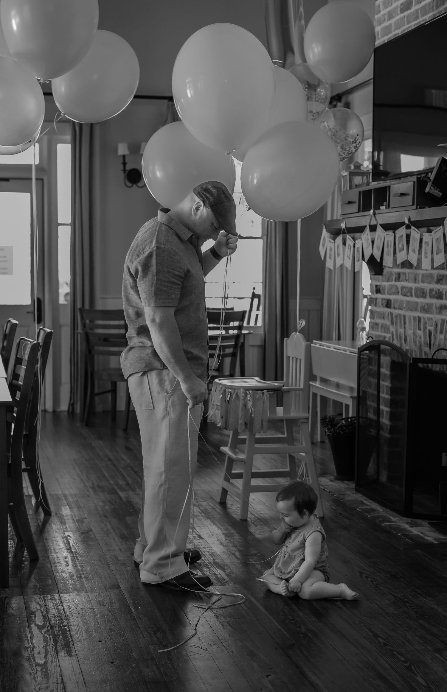 A black and white image showing a person holding balloons while a small child sits on a hardwood floor.