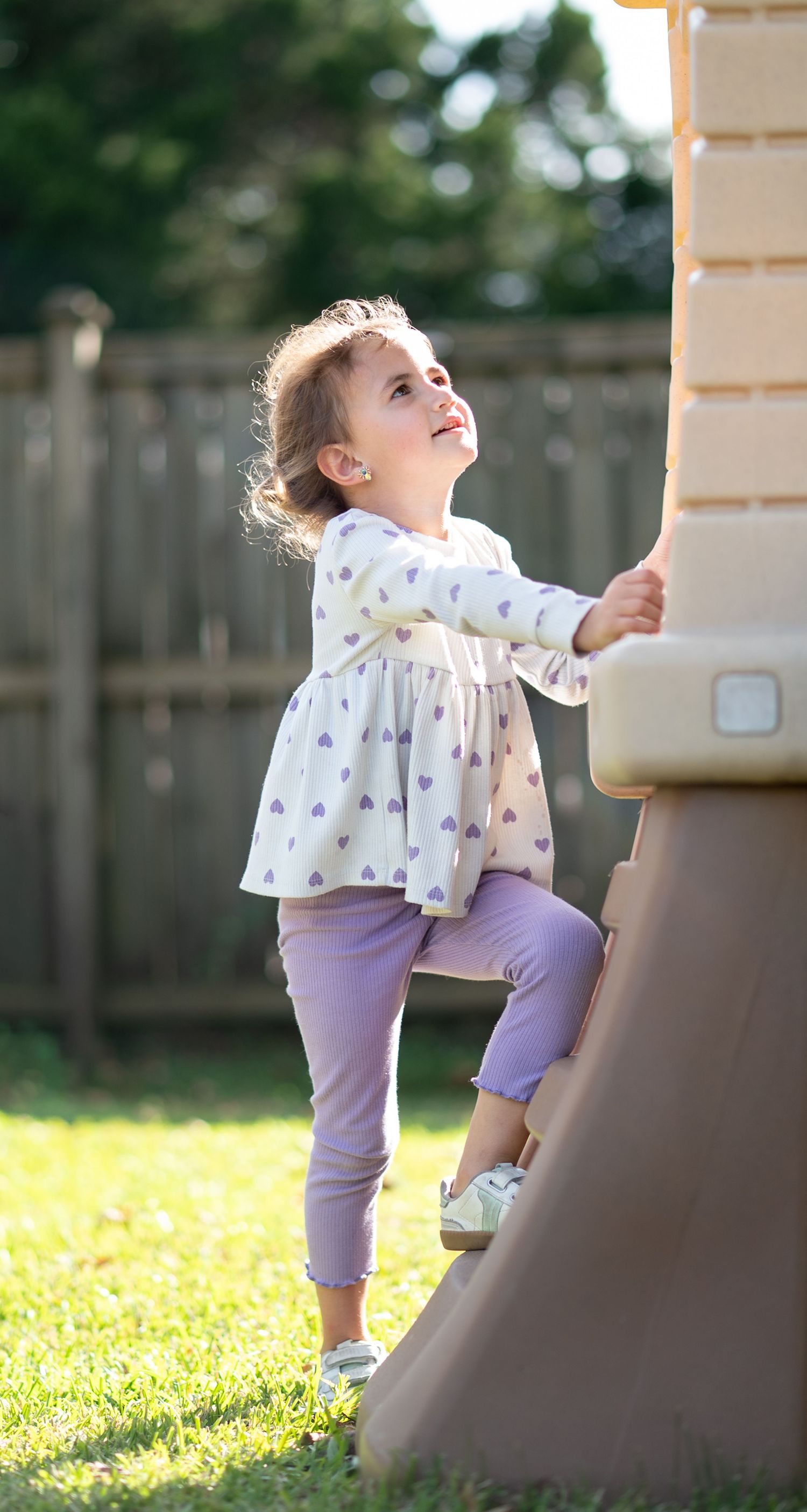 A toddler girl in purple pants and heart-print top climbs a plastic playground structure in a sunny backyard.