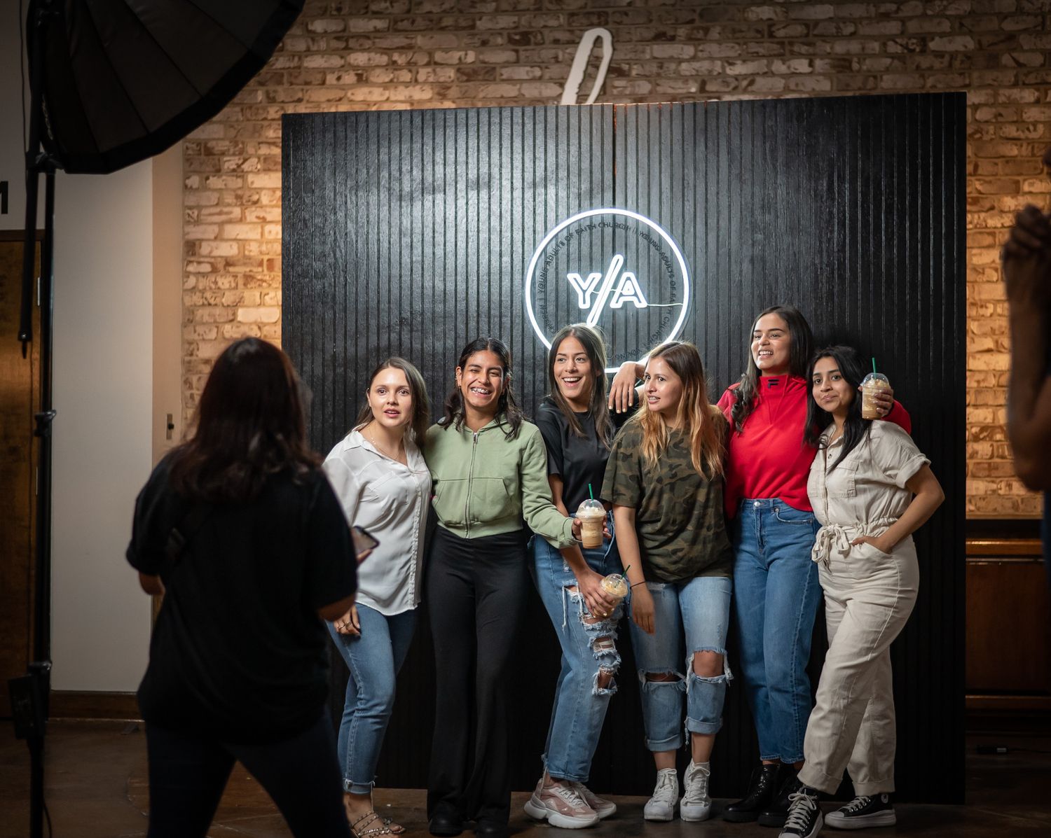 A group poses together against a brick wall with a neon sign in a casual indoor setting.