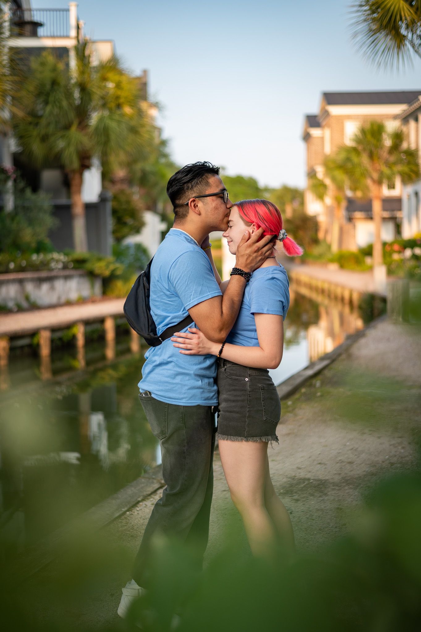 A couple embraces and kisses on a path beside a waterway with palm trees and buildings in the background.
