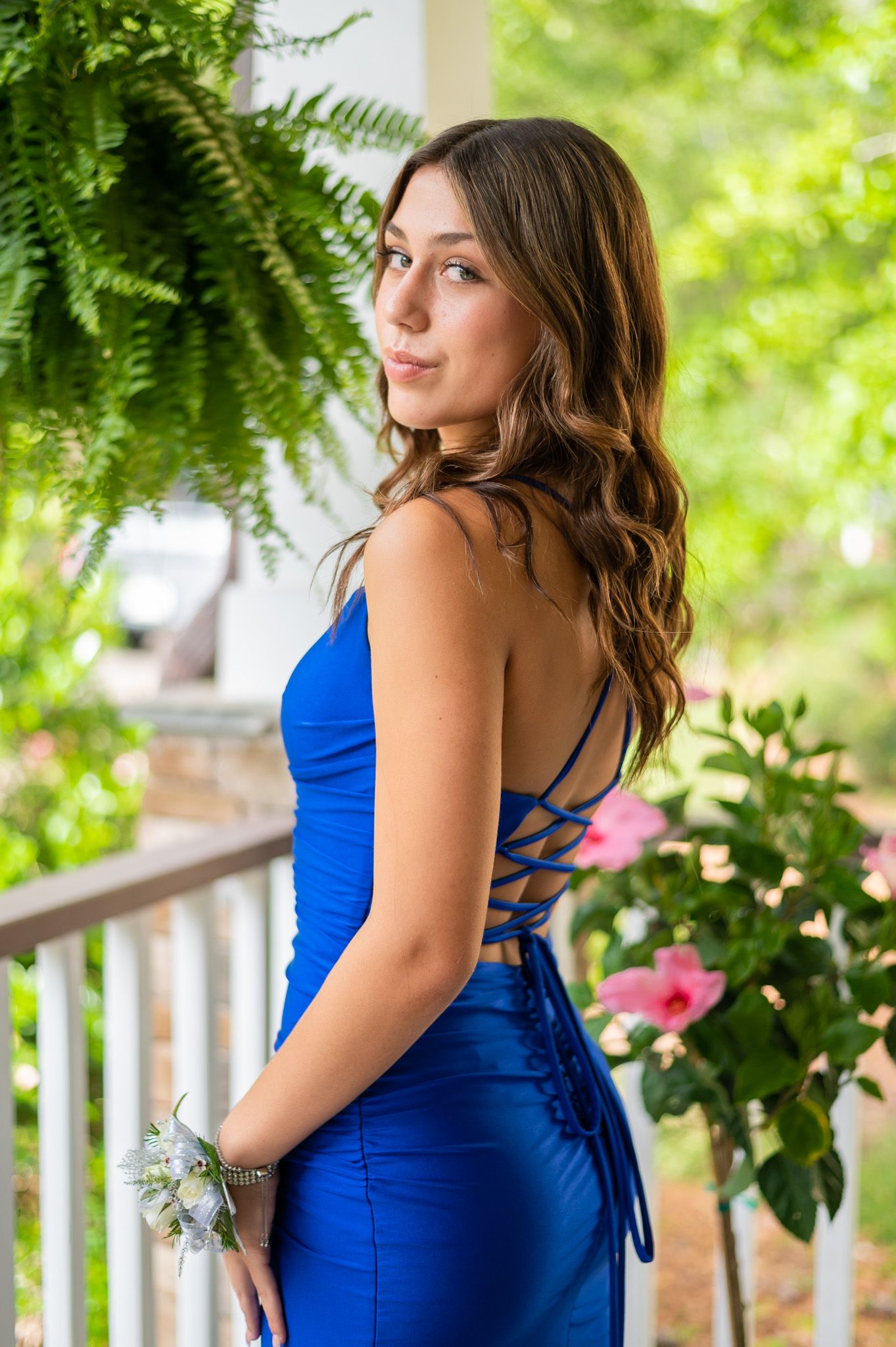 A woman in a vibrant blue dress poses on a porch with lush greenery and white railings in the background.
