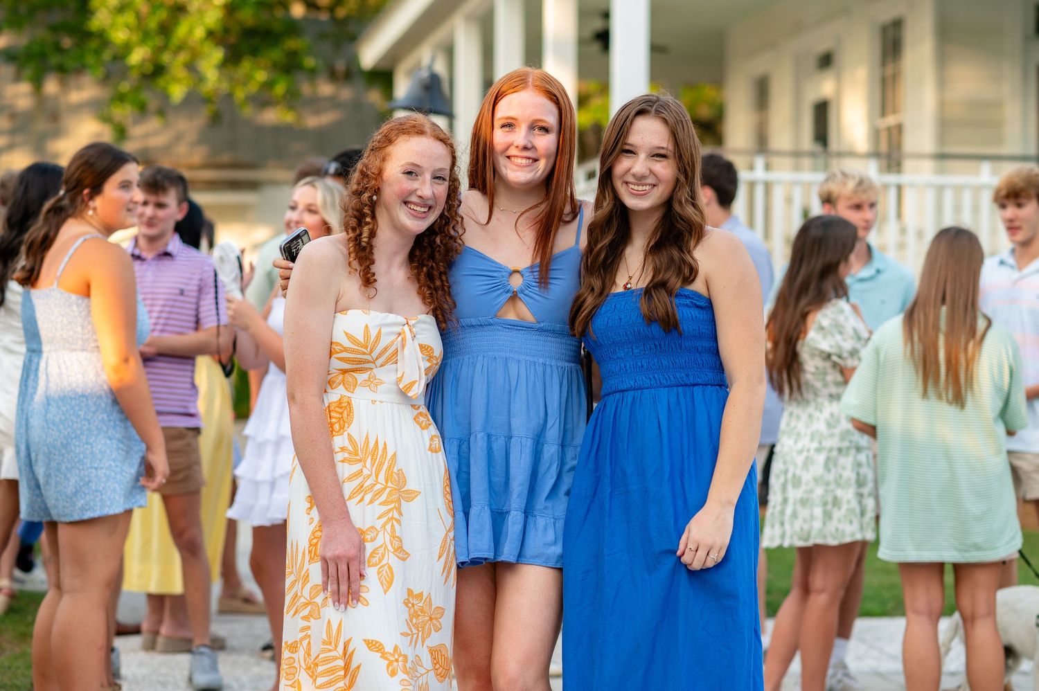 Three friends in summer dresses smile together at an outdoor social gathering.