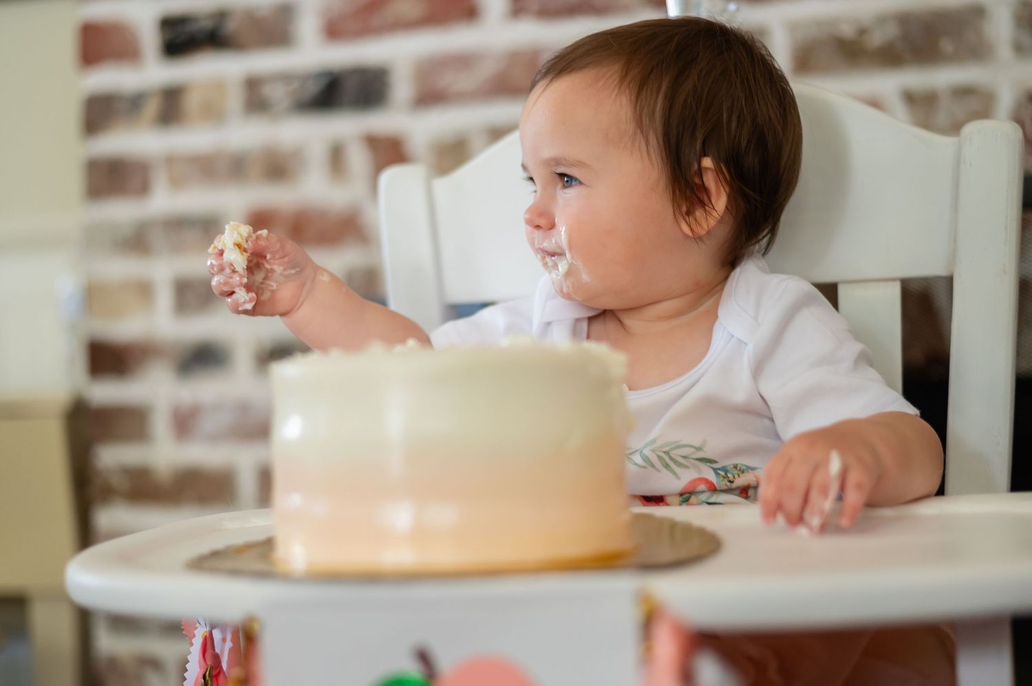 A young child sits in a high chair at mealtime with a cake on the table in front of a brick wall.
