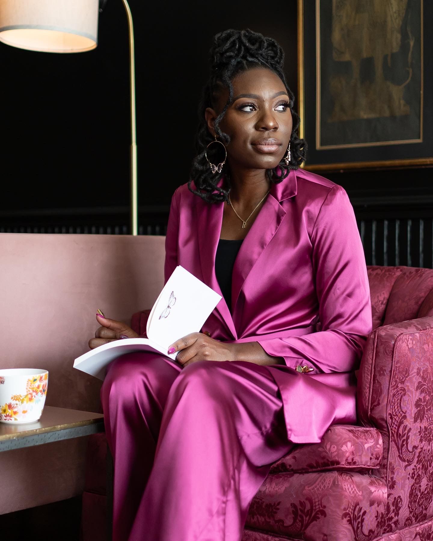 A person in a vibrant pink silk robe sits elegantly on a luxurious velvet couch in a dimly lit room.