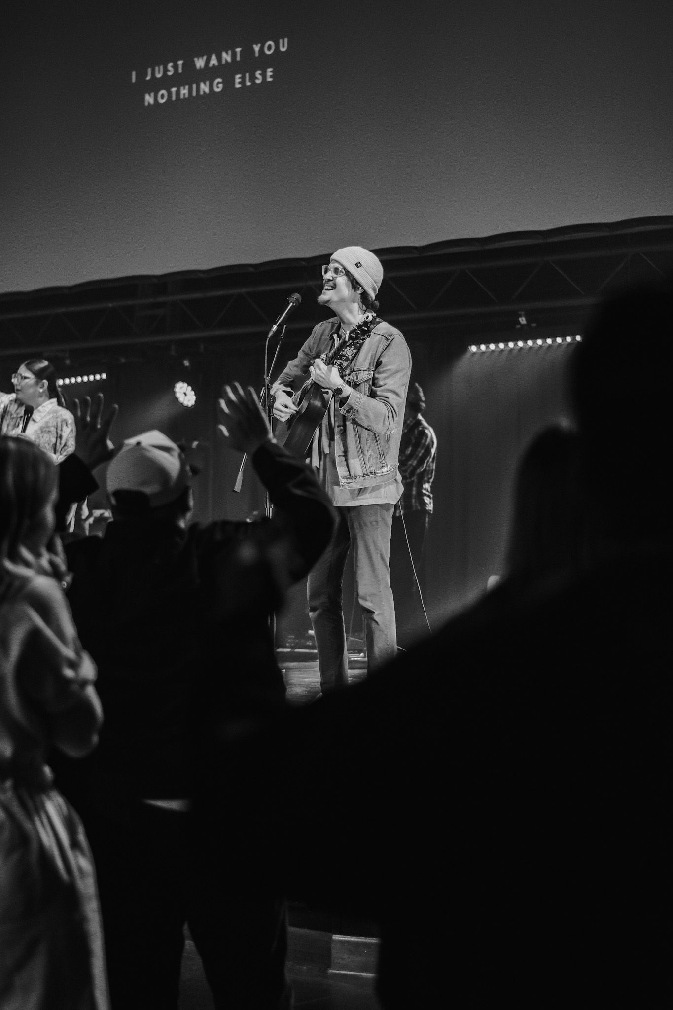 A performer in a denim jacket sings and plays guitar on stage during a black and white concert performance.