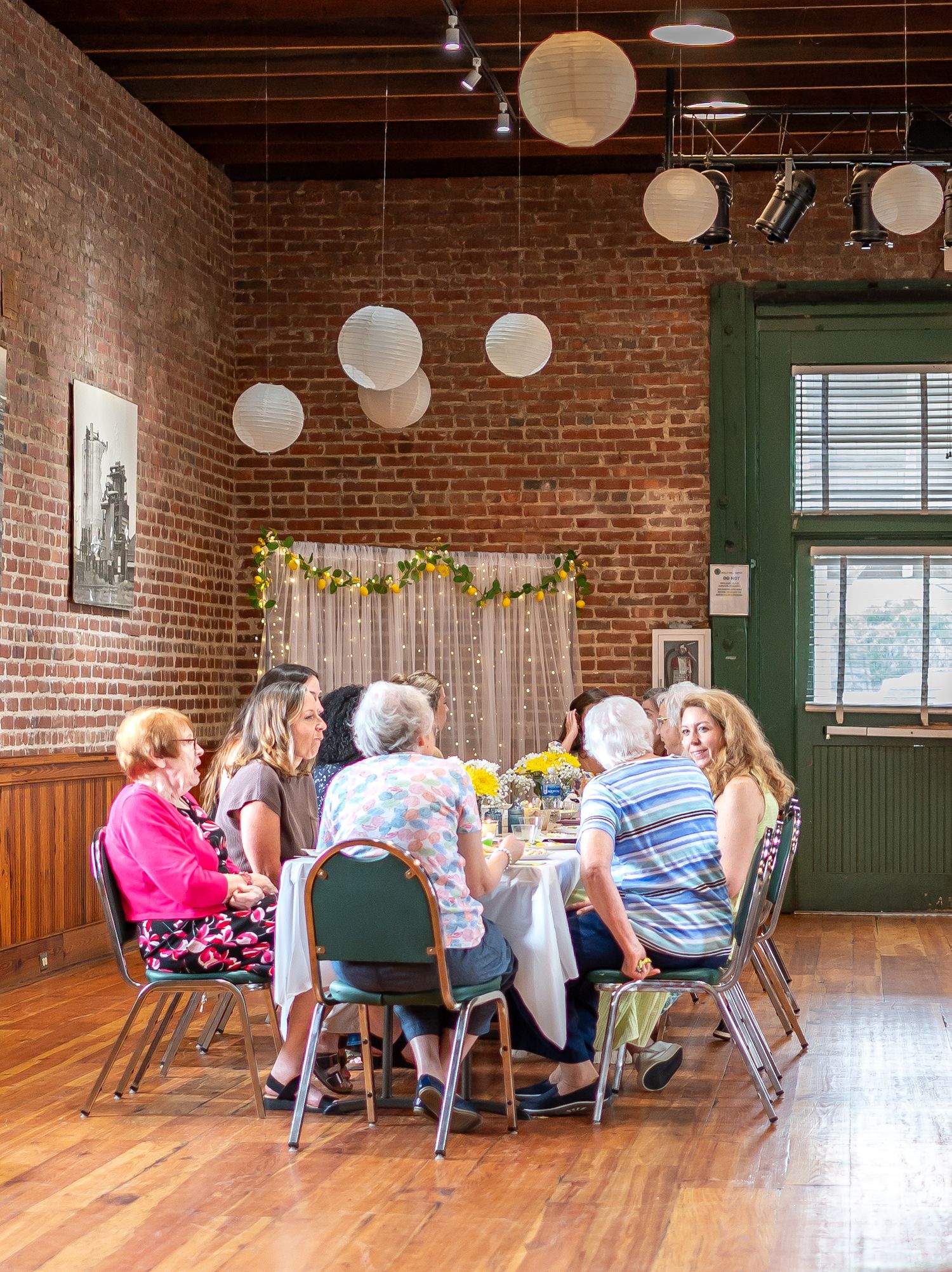 People gathered around tables in a rustic brick-walled event space with paper lanterns hanging from wooden beams.