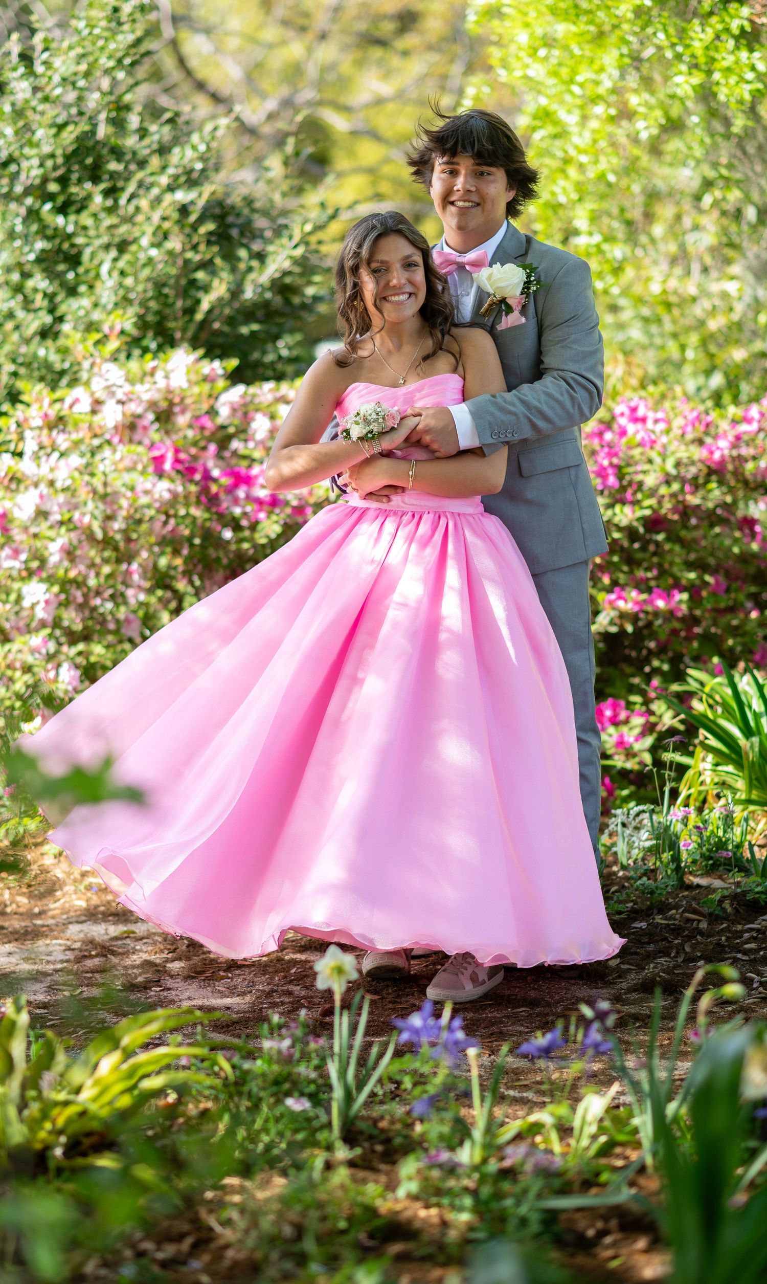 A couple poses romantically in a garden with blooming pink azaleas while the person in a pink ball gown twirls.
