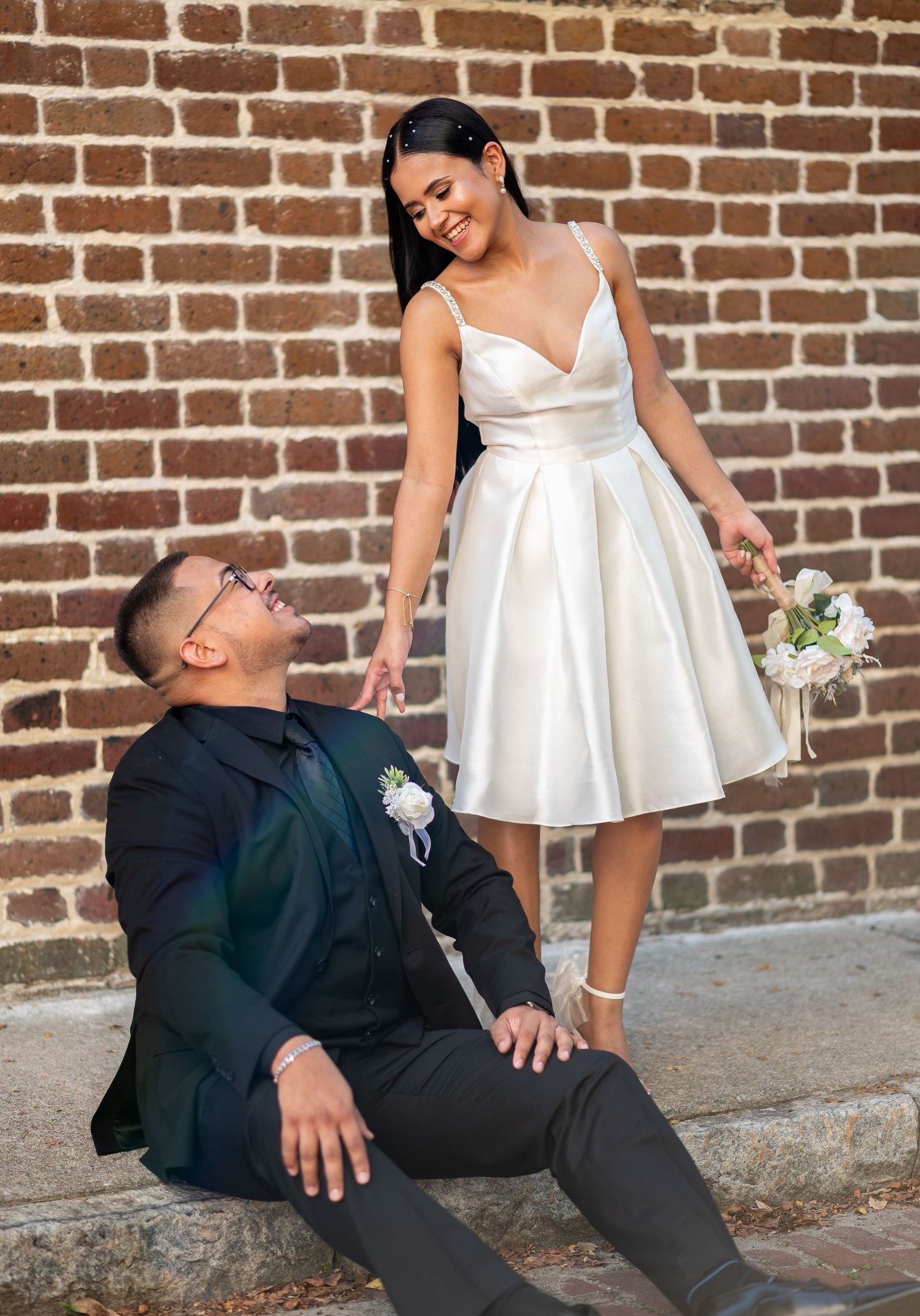 Couple posing playfully against brick wall during wedding photoshoot.