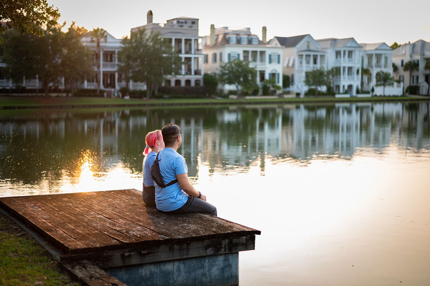 Couple sitting on wooden dock by tranquil lake at sunset creating romantic silhouette against golden sky.