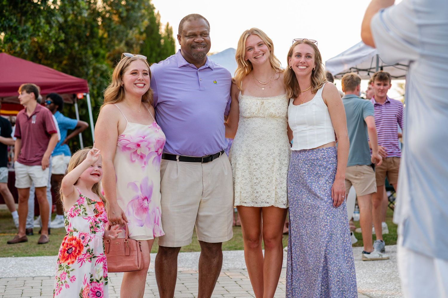Group of people gathered outdoors in summer attire at a social event with tents visible in background.