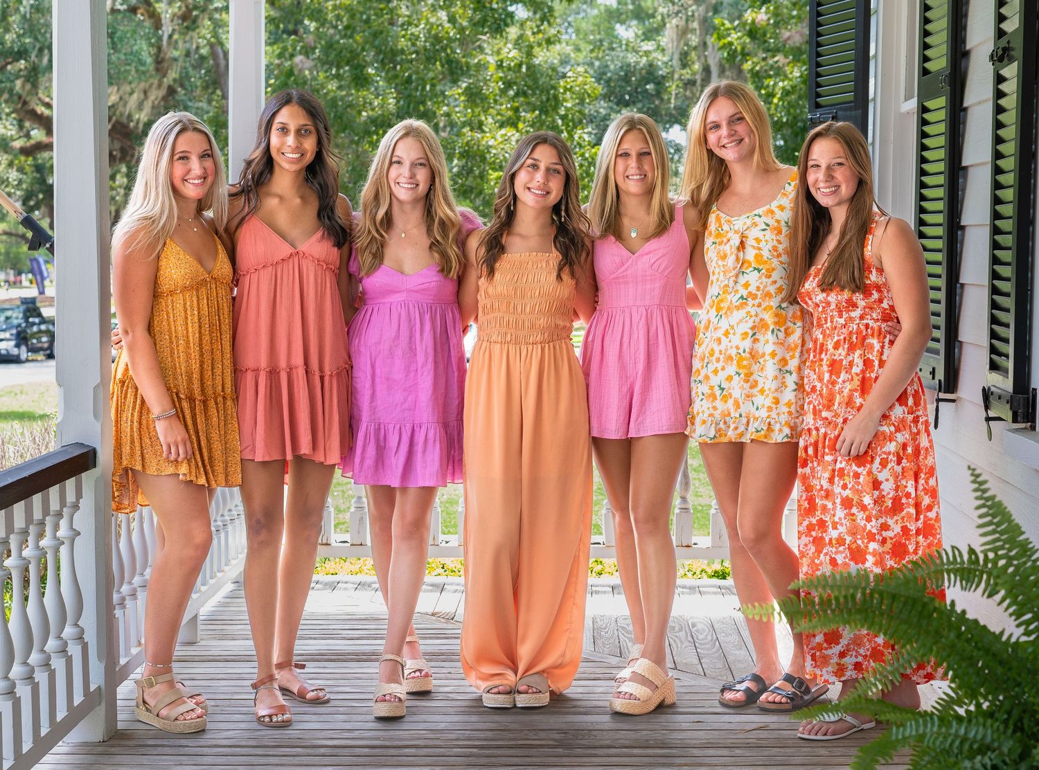A group of women in colorful summer dresses pose together on a white porch for a special occasion.