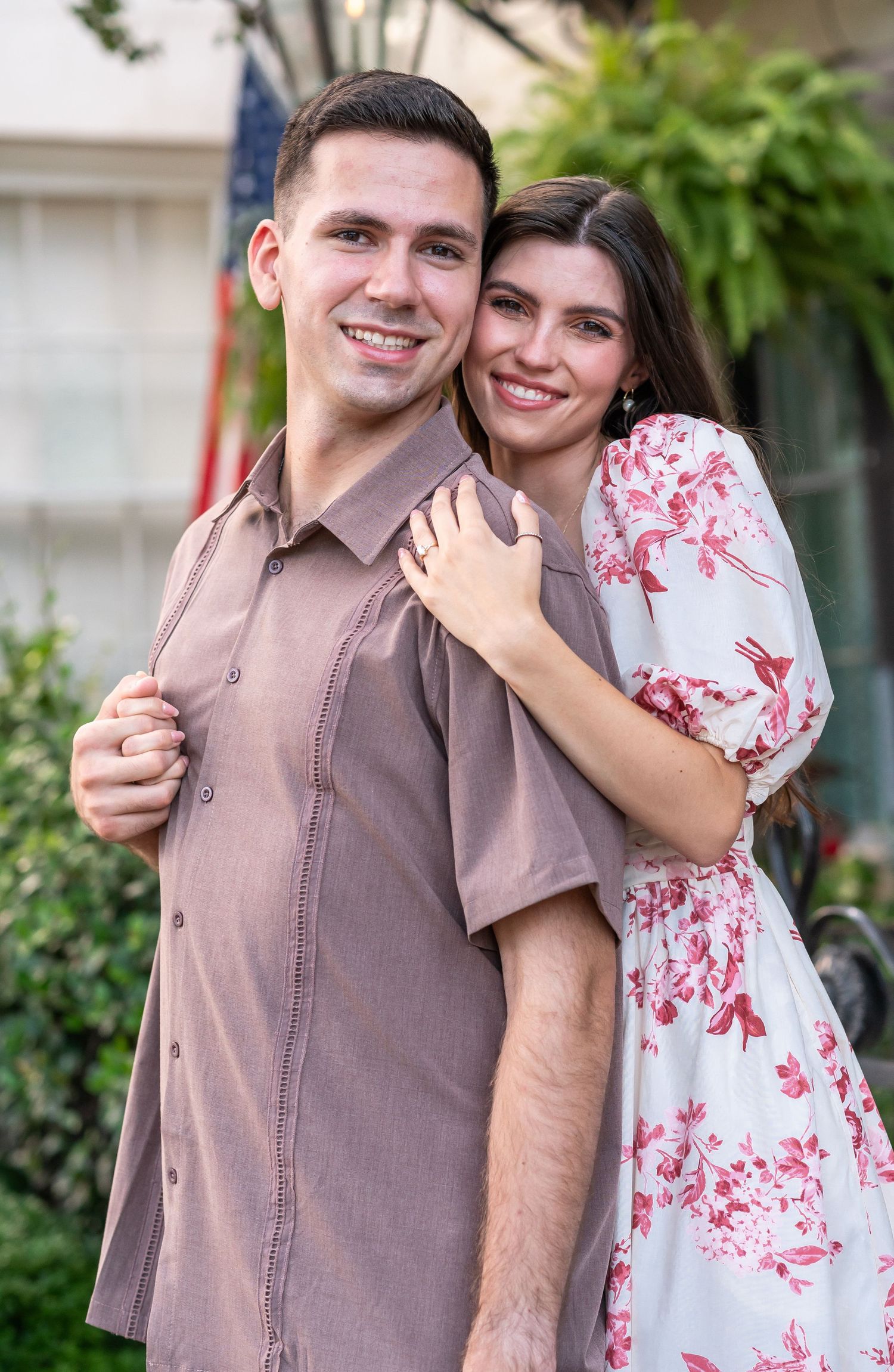 A couple poses together outdoors, with her wearing a white and red floral dress and him in a brown button-down shirt.