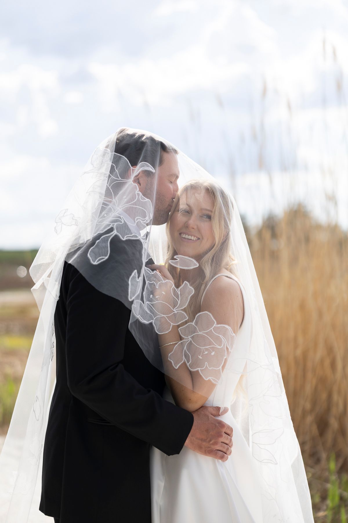 Bride and groom share a romantic embrace under a floral lace veil outdoors on their wedding day.