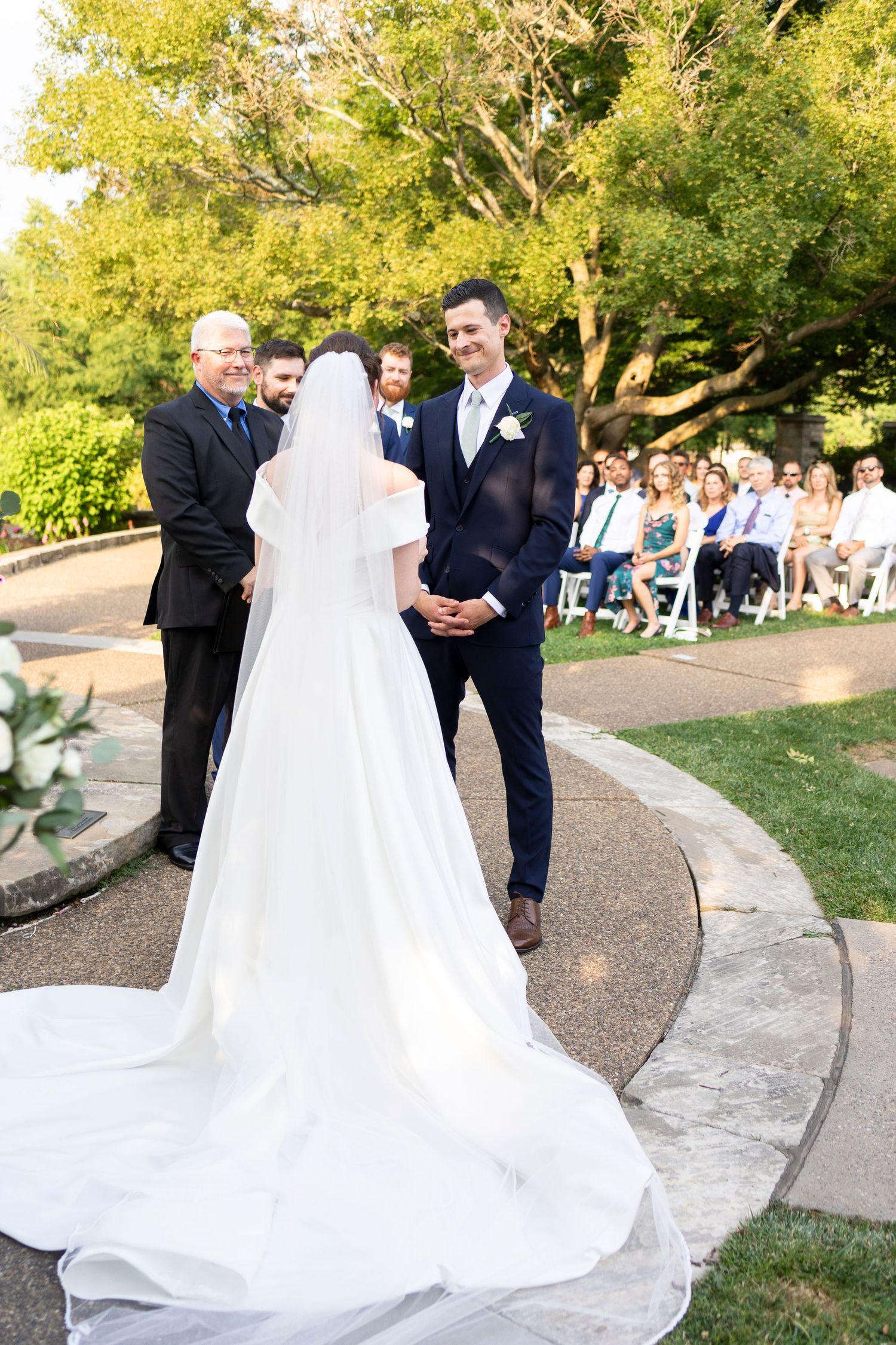 Wedding ceremony in progress with long train of bride's dress visible on curved garden path.