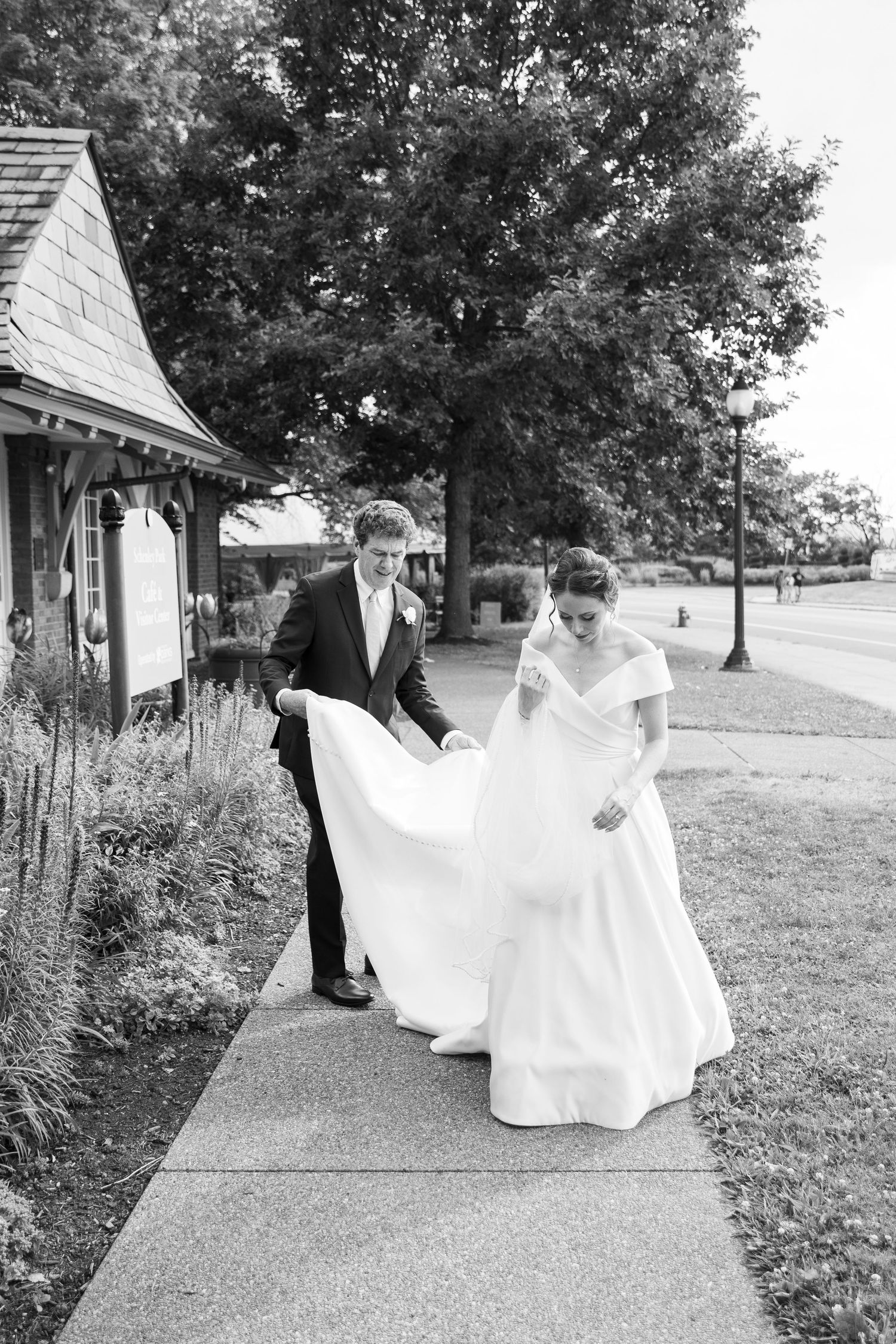 A couple in wedding attire walks along a garden path in black and white photography.