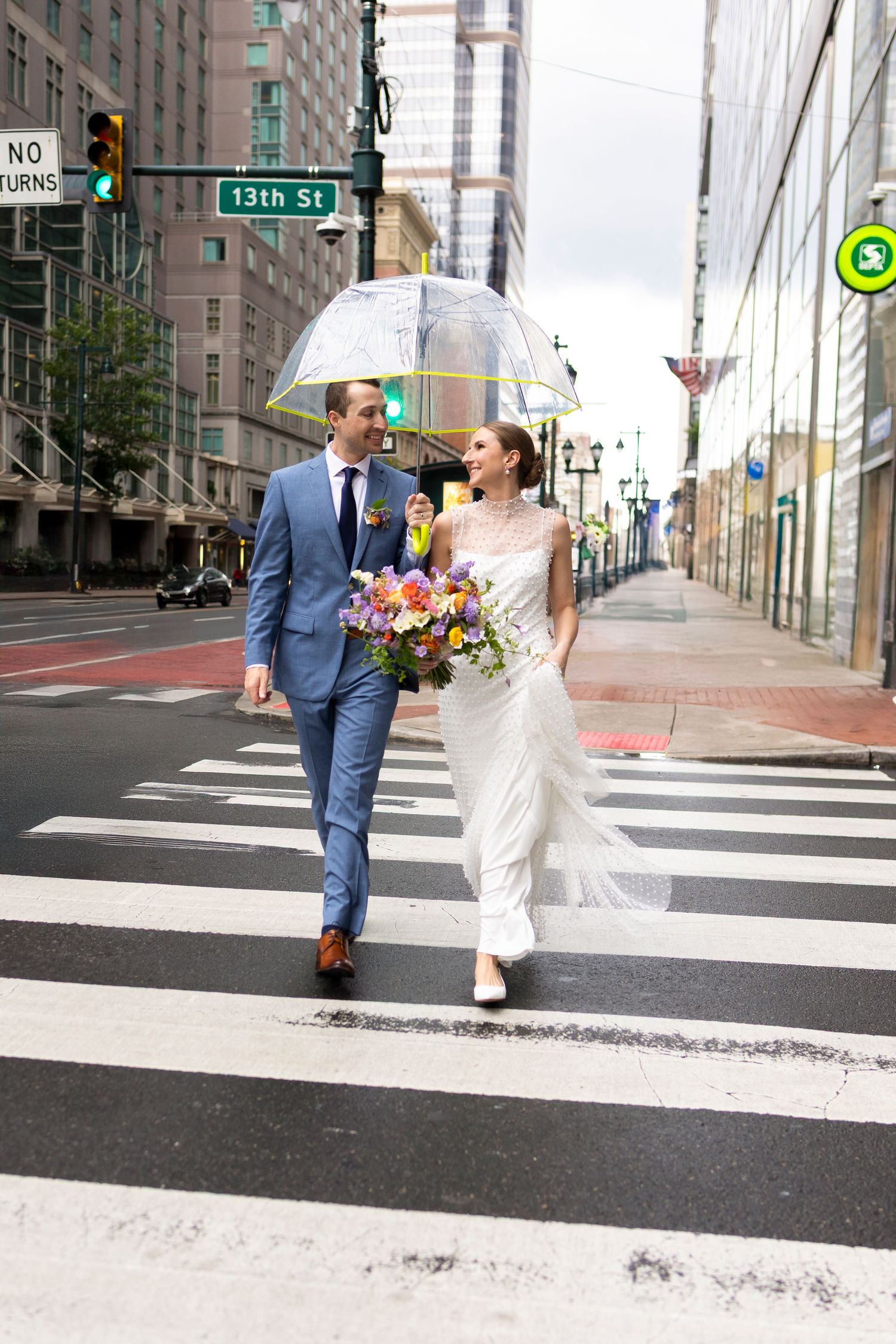 Newlyweds cross urban street with clear umbrella and colorful bouquet at 13th Street intersection.