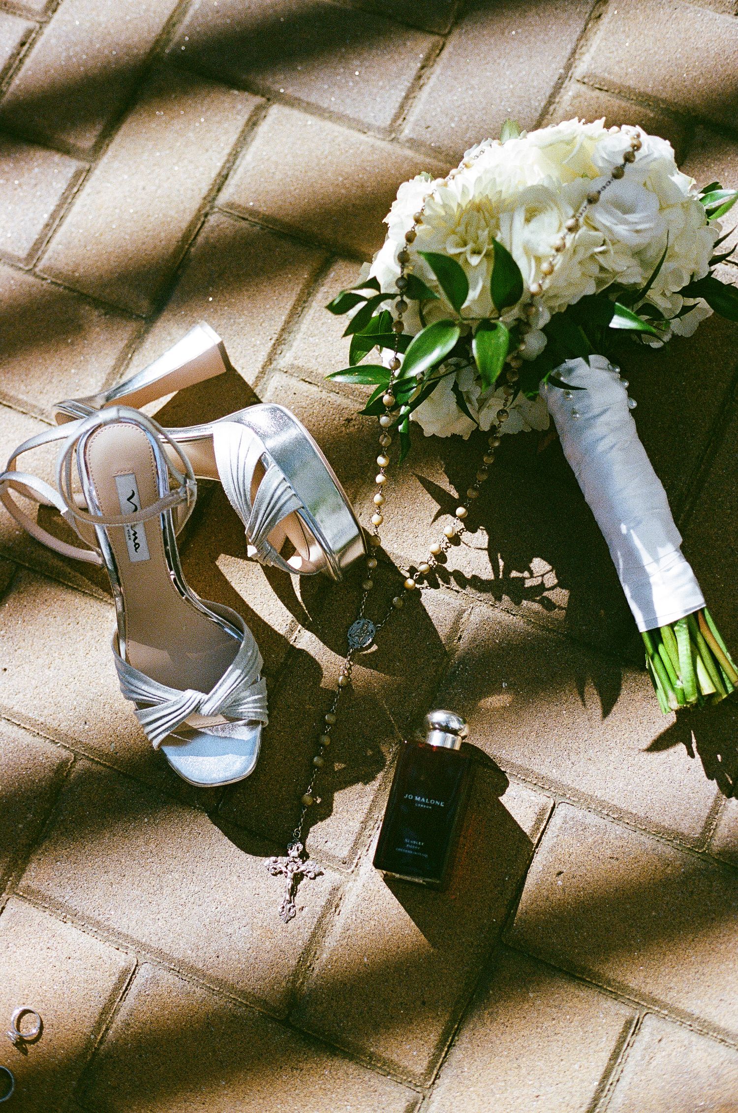 Wedding accessories including silver shoes, white bouquet and rings arranged on tiled floor.