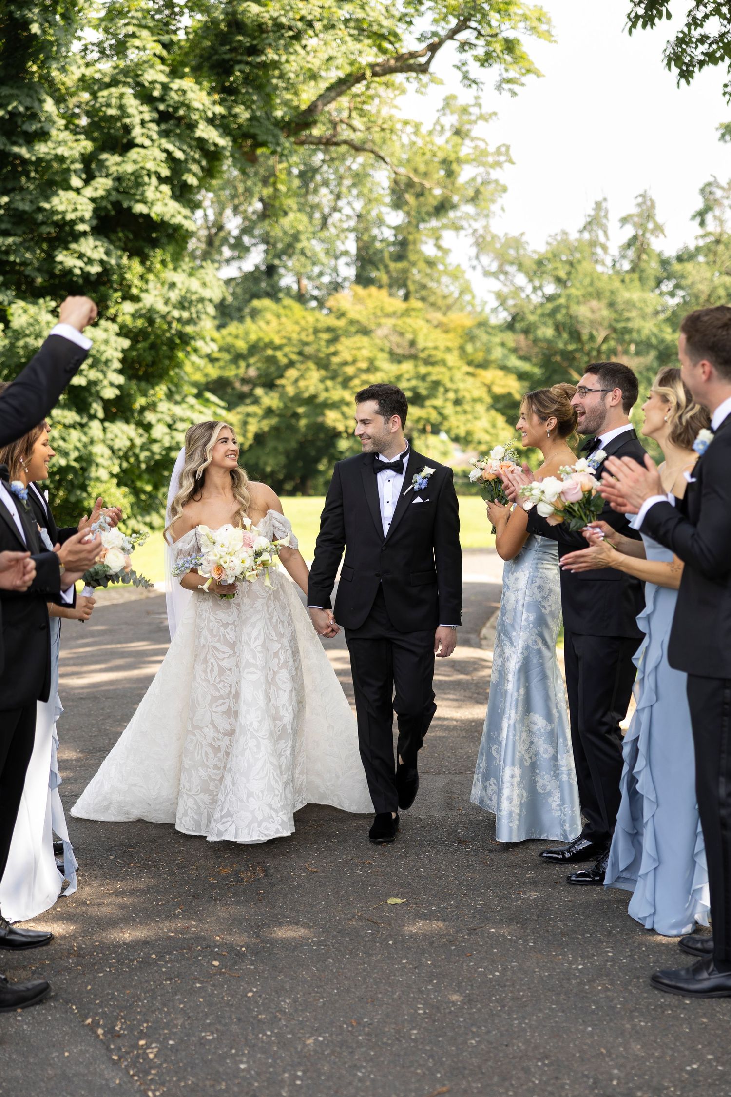 Wedding party in light blue dresses and black tuxedos walking and laughing outdoors.