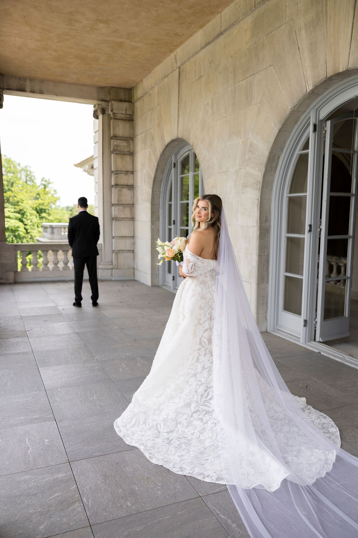 First look wedding photos showing a bride in a lace gown with long train on an elegant stone balcony.