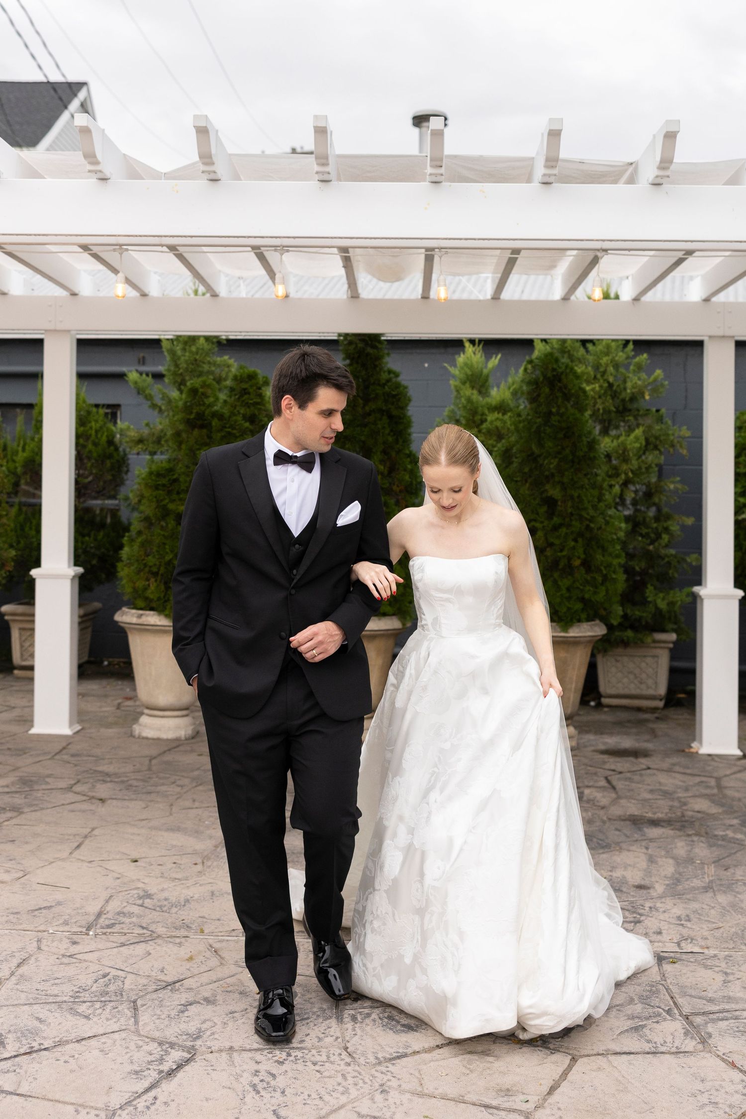 A couple shares a tender moment while walking under a white pergola with stone pavers.