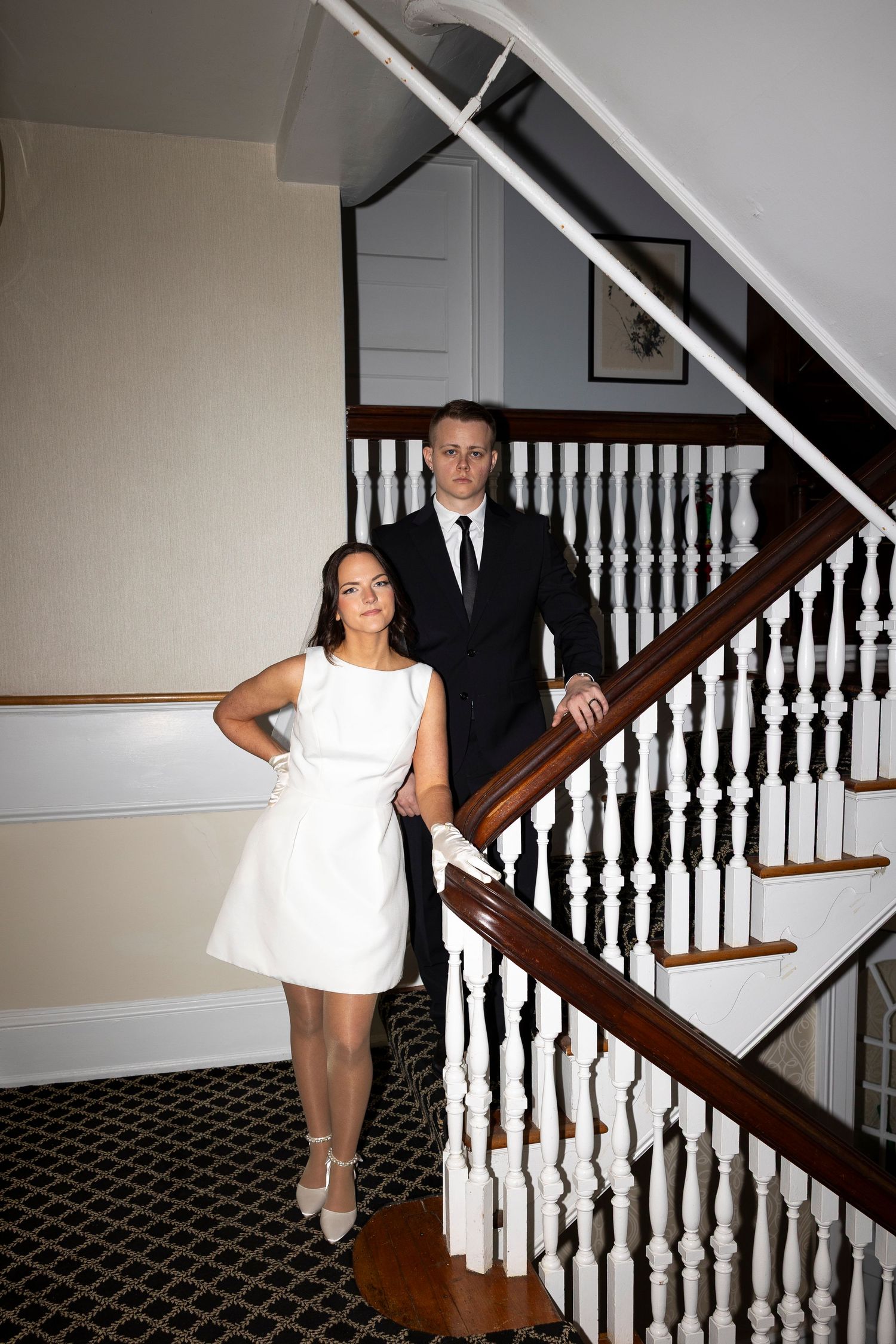 A couple poses on a classic white-bannistered staircase in formal attire.