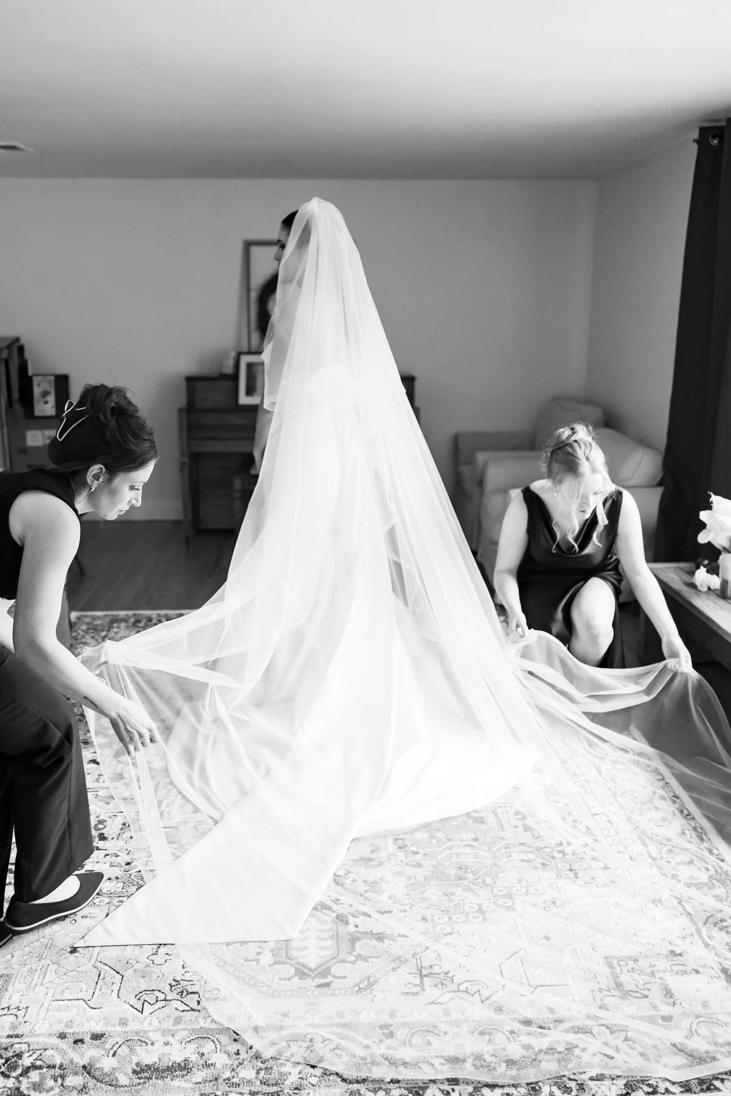 Two women in black dresses arranging a bride's long cathedral veil in a room, captured in black and white.