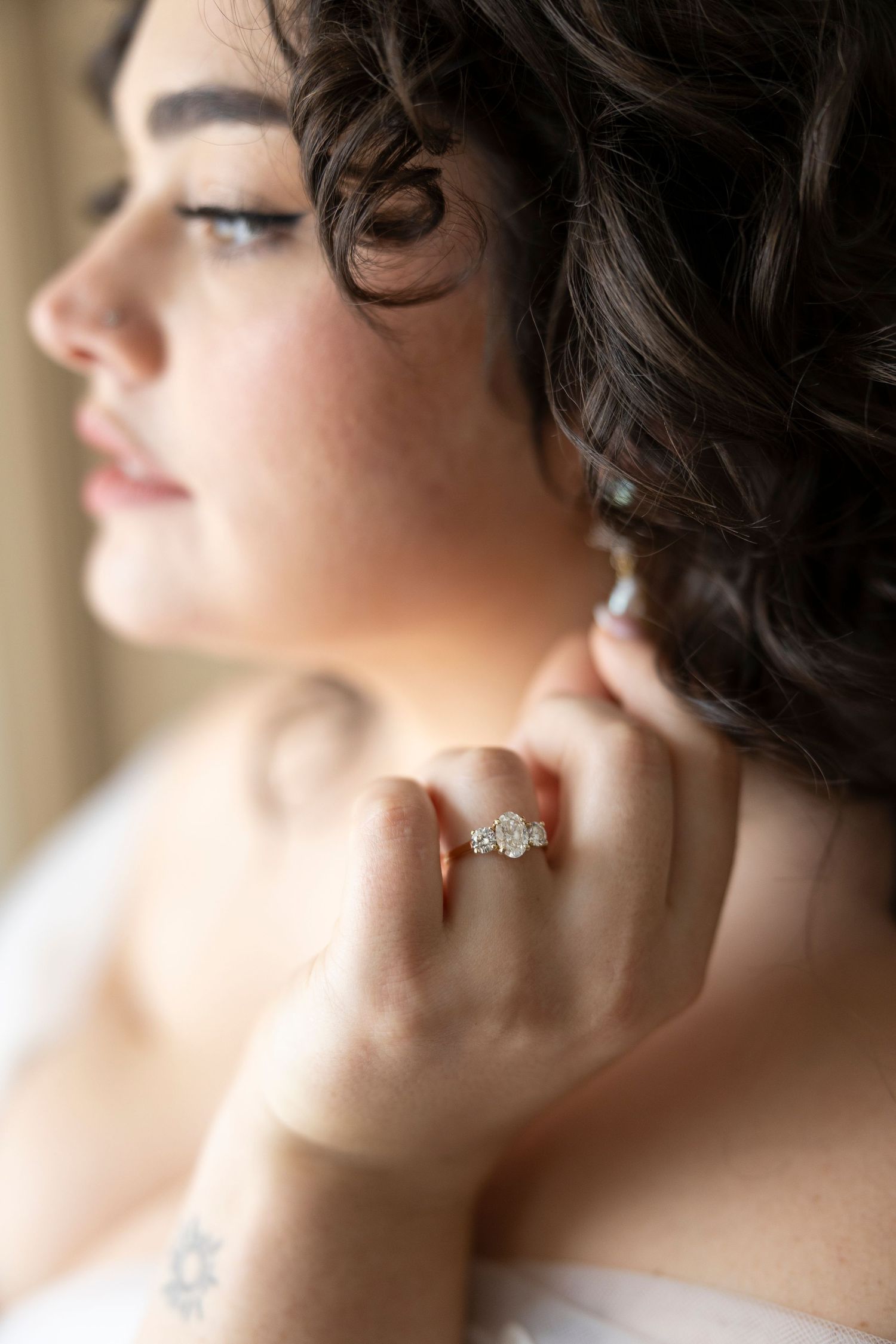 Bride adjusting earring with diamond engagement ring visible, dark curly hair, soft natural light, elegant bridal portrait.