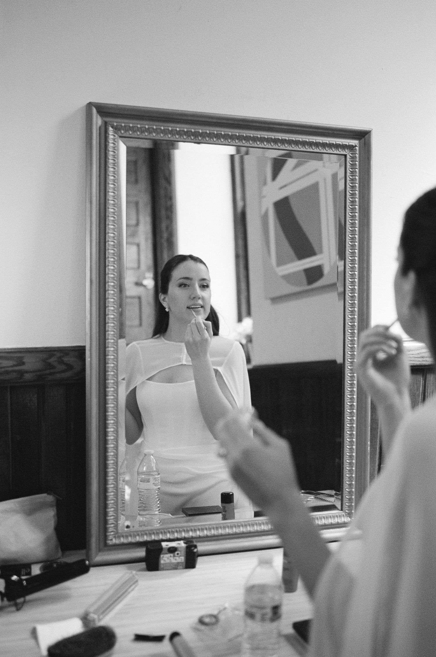 Black and white photo of a bride applying makeup at a vanity mirror in a white dress.