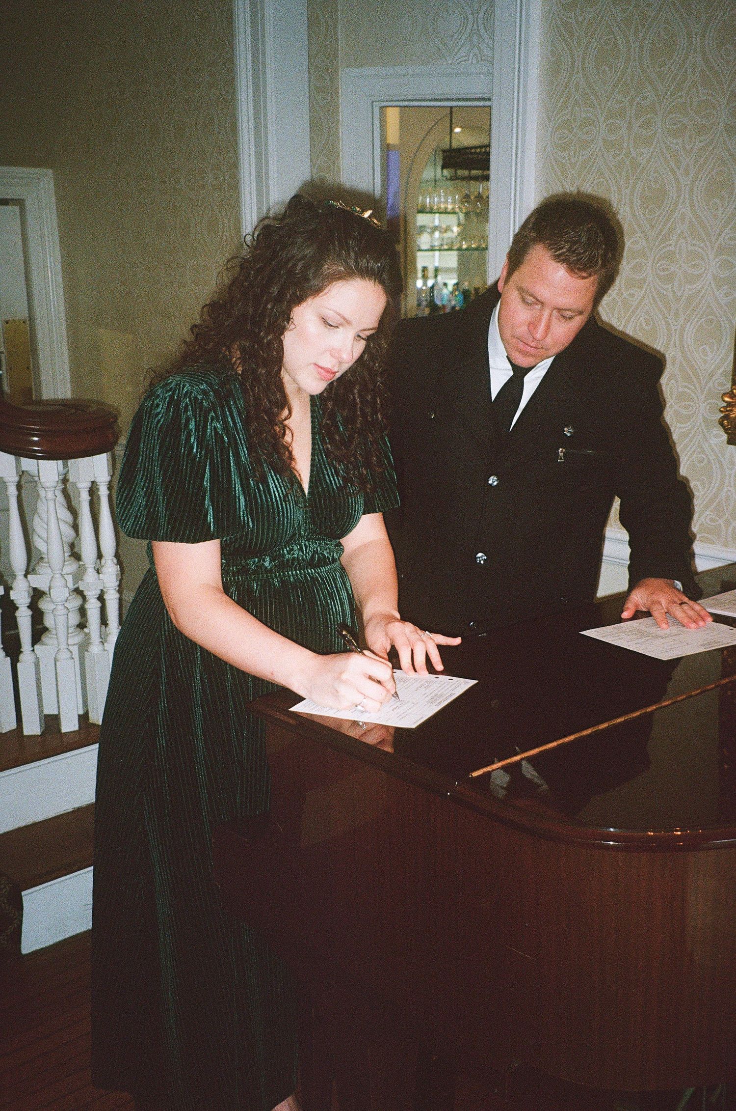 Two people in formal attire sit at a piano reviewing documents.