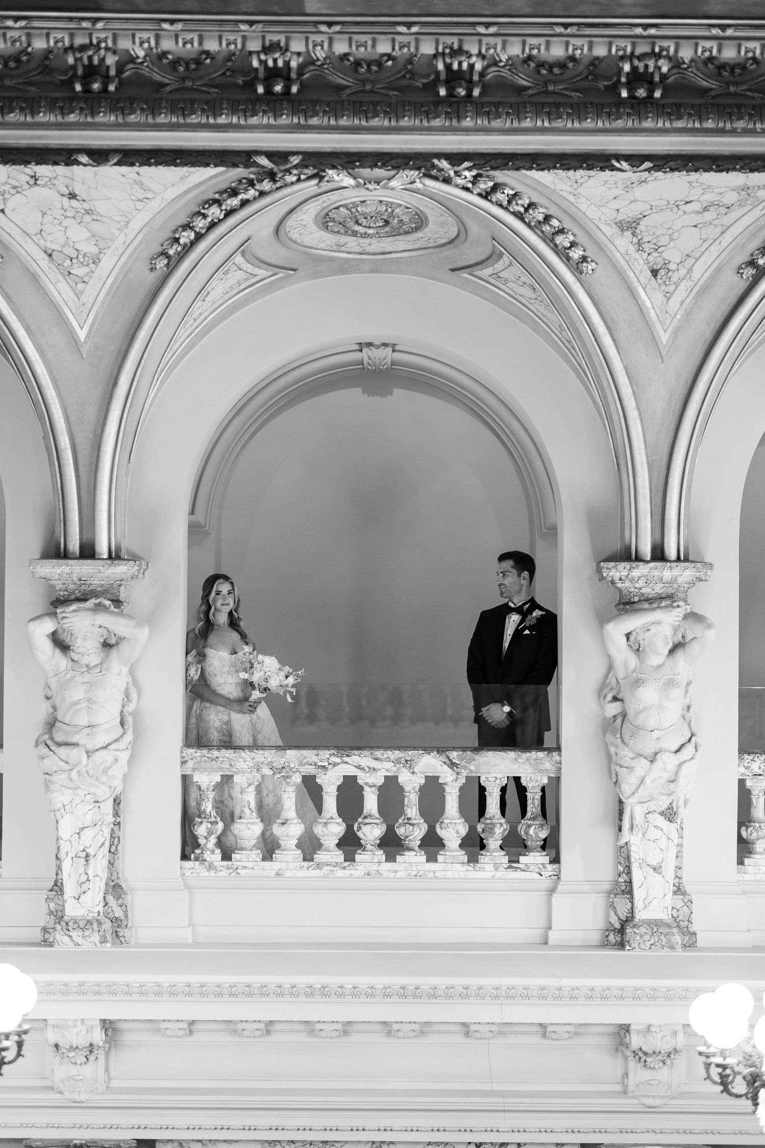 Elegant wedding couple poses in ornate white architectural balcony with decorative moldings and columns.