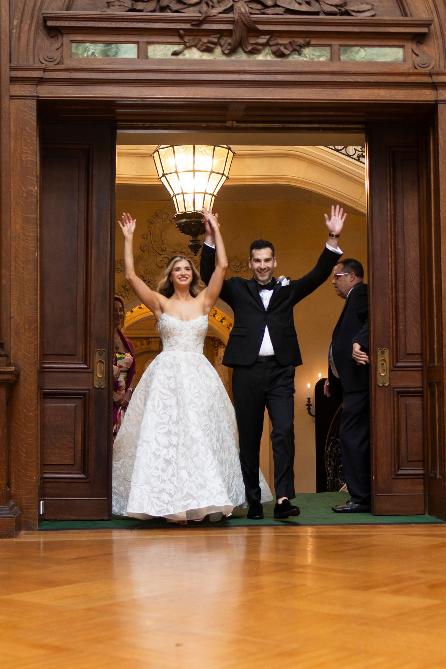 Wedding party making grand entrance through ornate wooden doorway with raised arms.