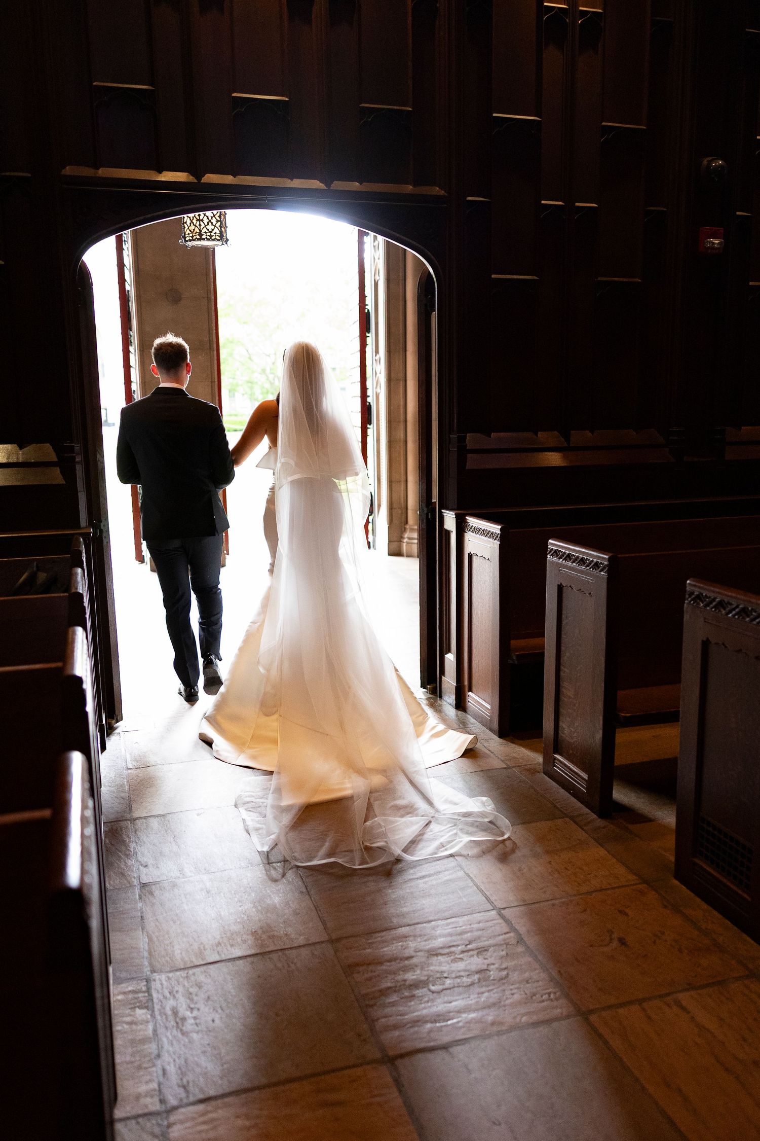 Bride and groom walking hand in hand toward church doors, with wedding dress train and veil trailing on stone floor.